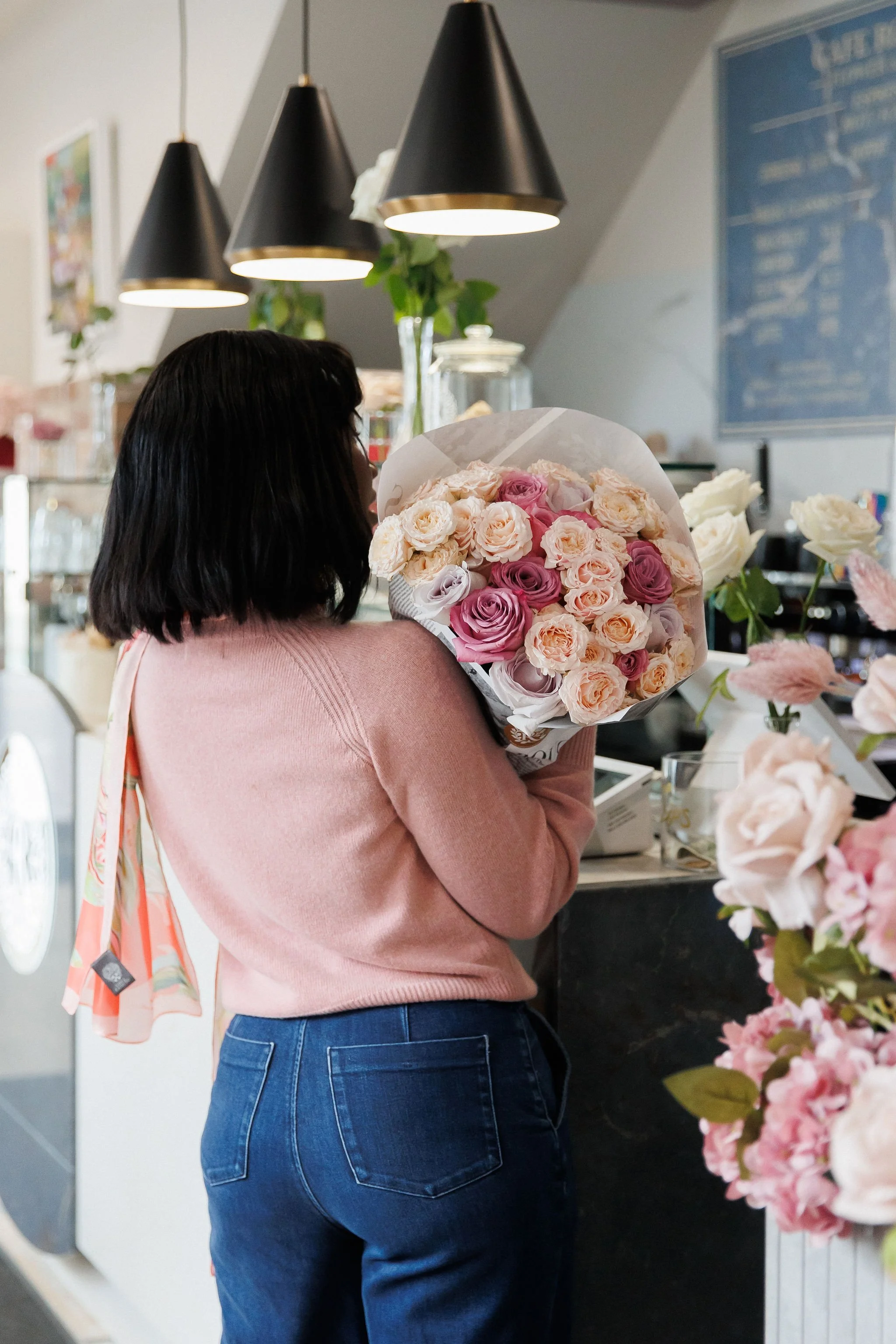 A woman with dark hair in a pink sweater and blue jeans standing at a counter in a flower shop, holding a bouquet of pink, purple, and cream roses.