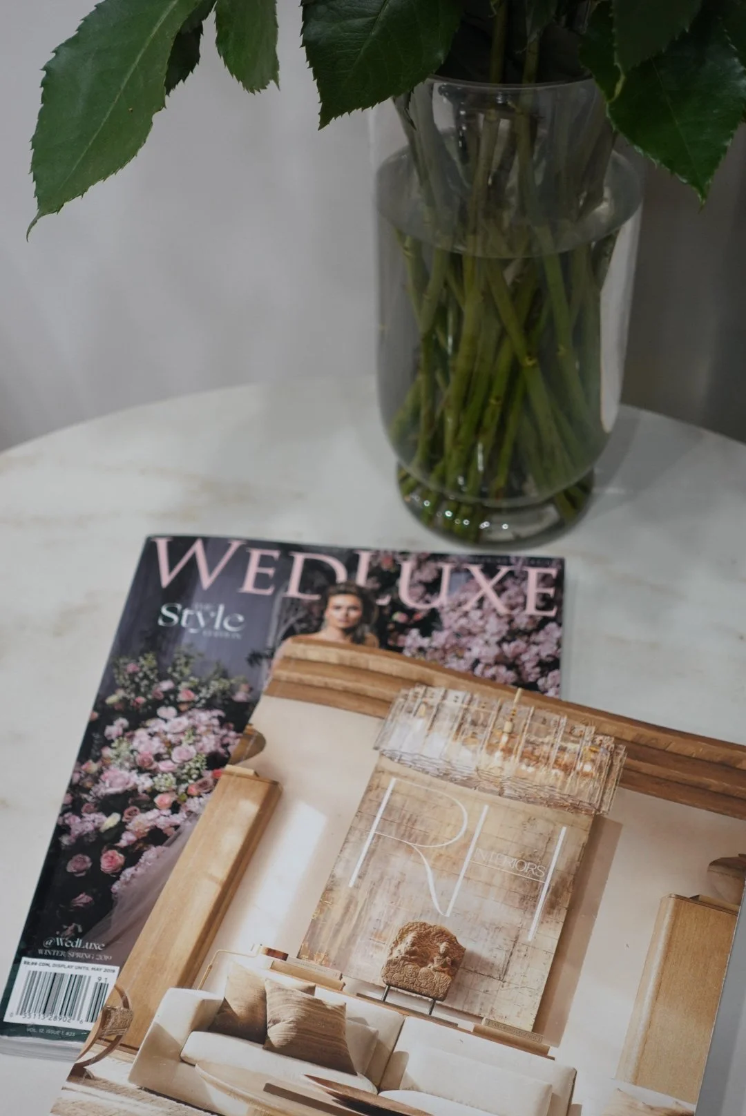 A glass vase with green leaves and stems, a copy of WEDLUXE magazine, and a wooden furniture catalog on a white marble table.