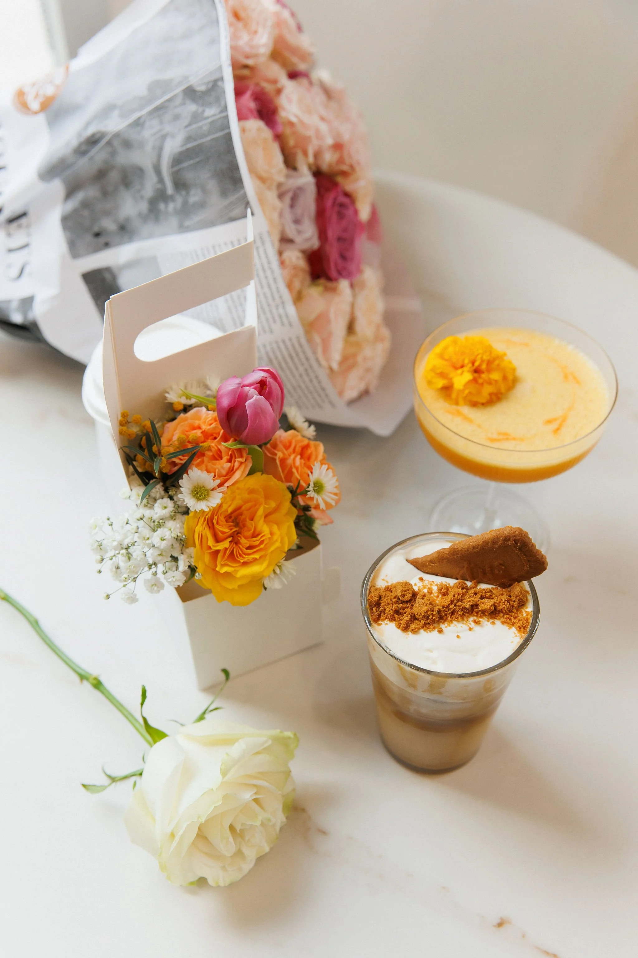 Various desserts and flowers on a white table, including a white rose, a small bouquet with tulips and daisies, a yellow flower in a glass, an orange-colored beverage with a yellow flower garnish, and a layered coffee drink with a cookie and crumb to