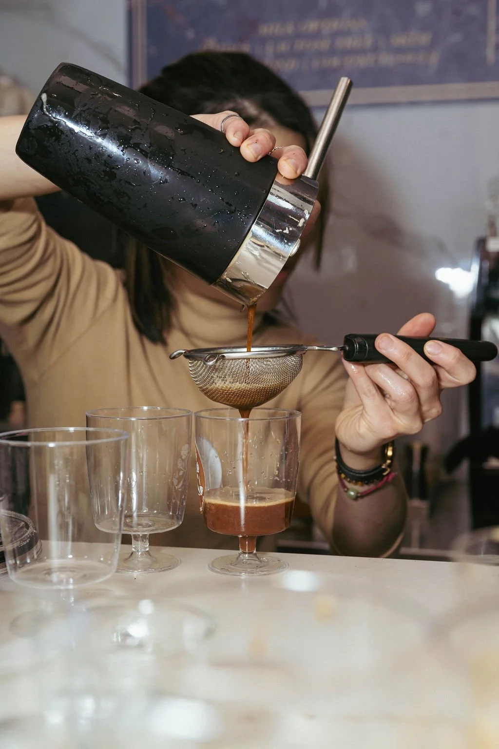 Person pouring coffee through a metal strainer into a glass, with several empty glasses on the table.