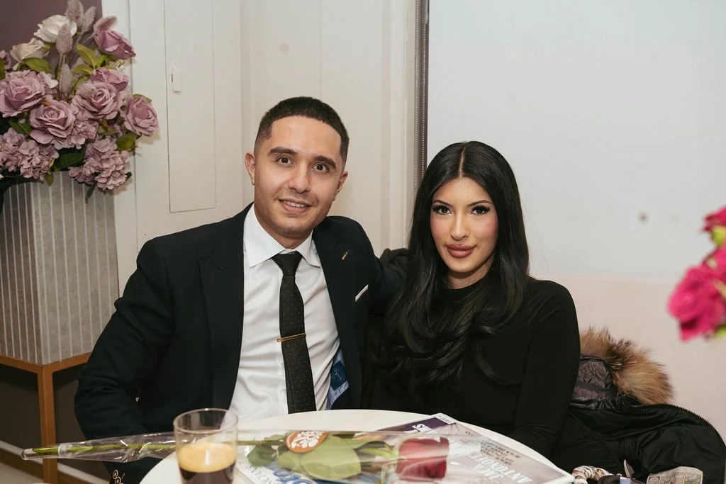 A man and woman sitting at a table in a restaurant or cafe, with flowers visible in the background.