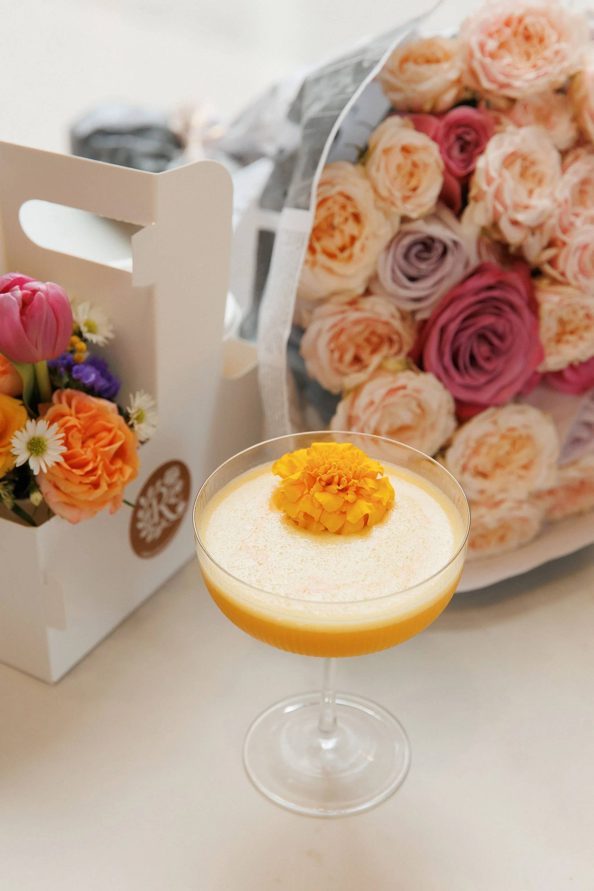 A cocktail with a yellow flower garnish, surrounded by bouquets of pink, purple, and peach roses and small mixed flowers.