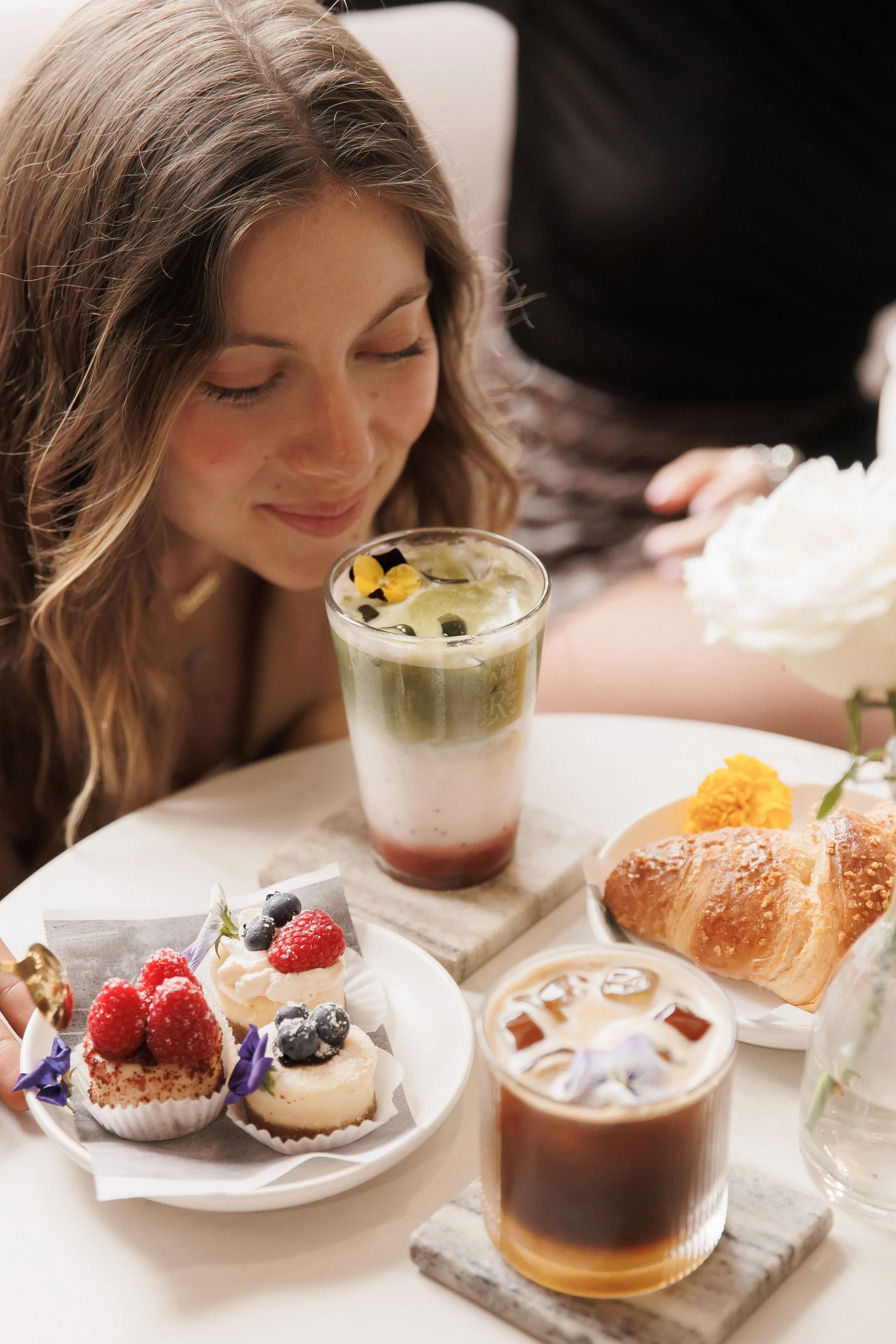 A woman with wavy light brown hair leaning towards a table with desserts and drinks, including a glass of layered cocktail with lime and berry garnishes, an iced coffee with ice cubes, a plate of assorted berry-topped cupcakes, a croissant, and a yel