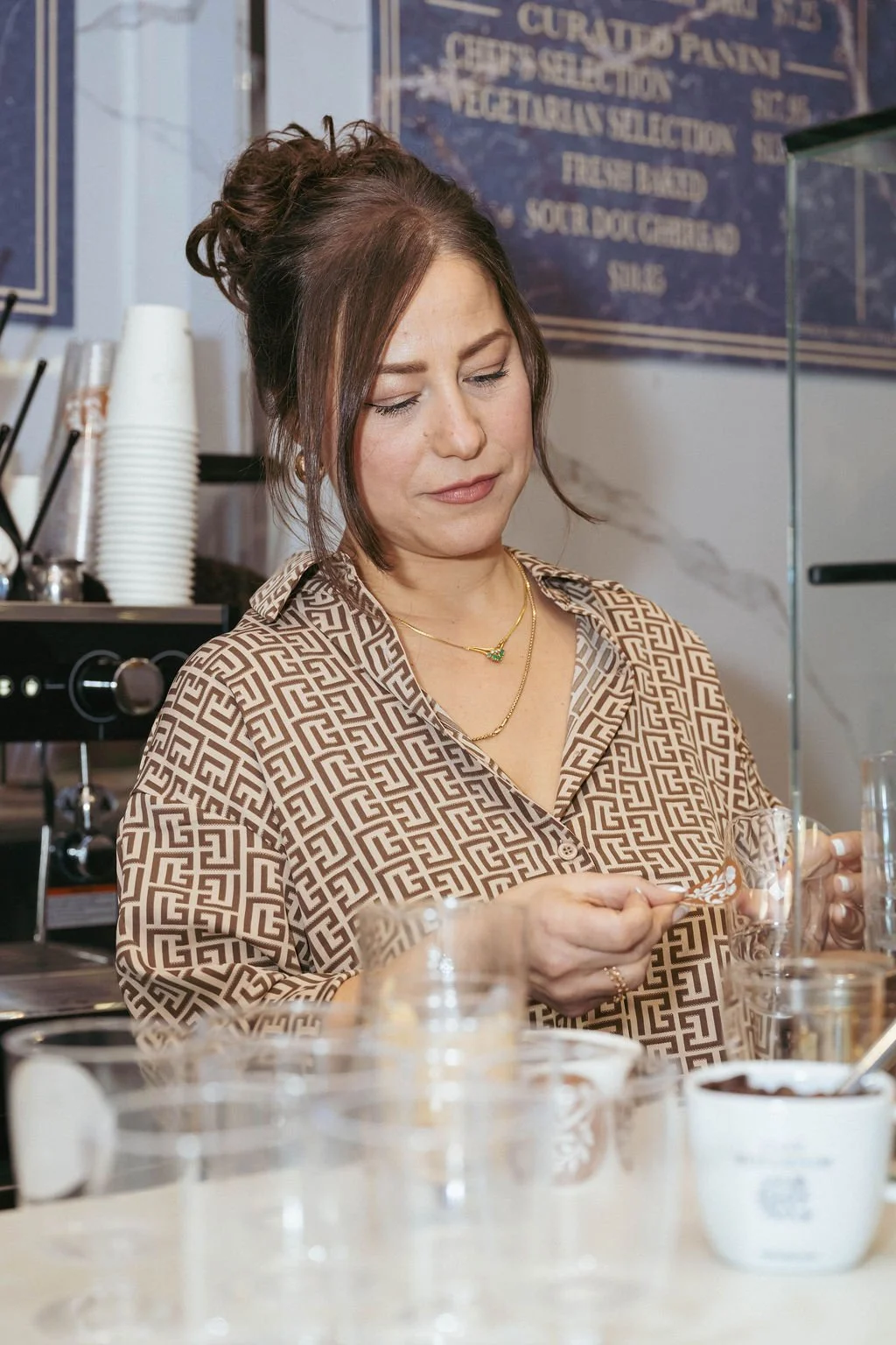 A woman with brown hair in a bun, wearing a patterned shirt and gold jewelry, is sitting at a cafe, looking at her phone.