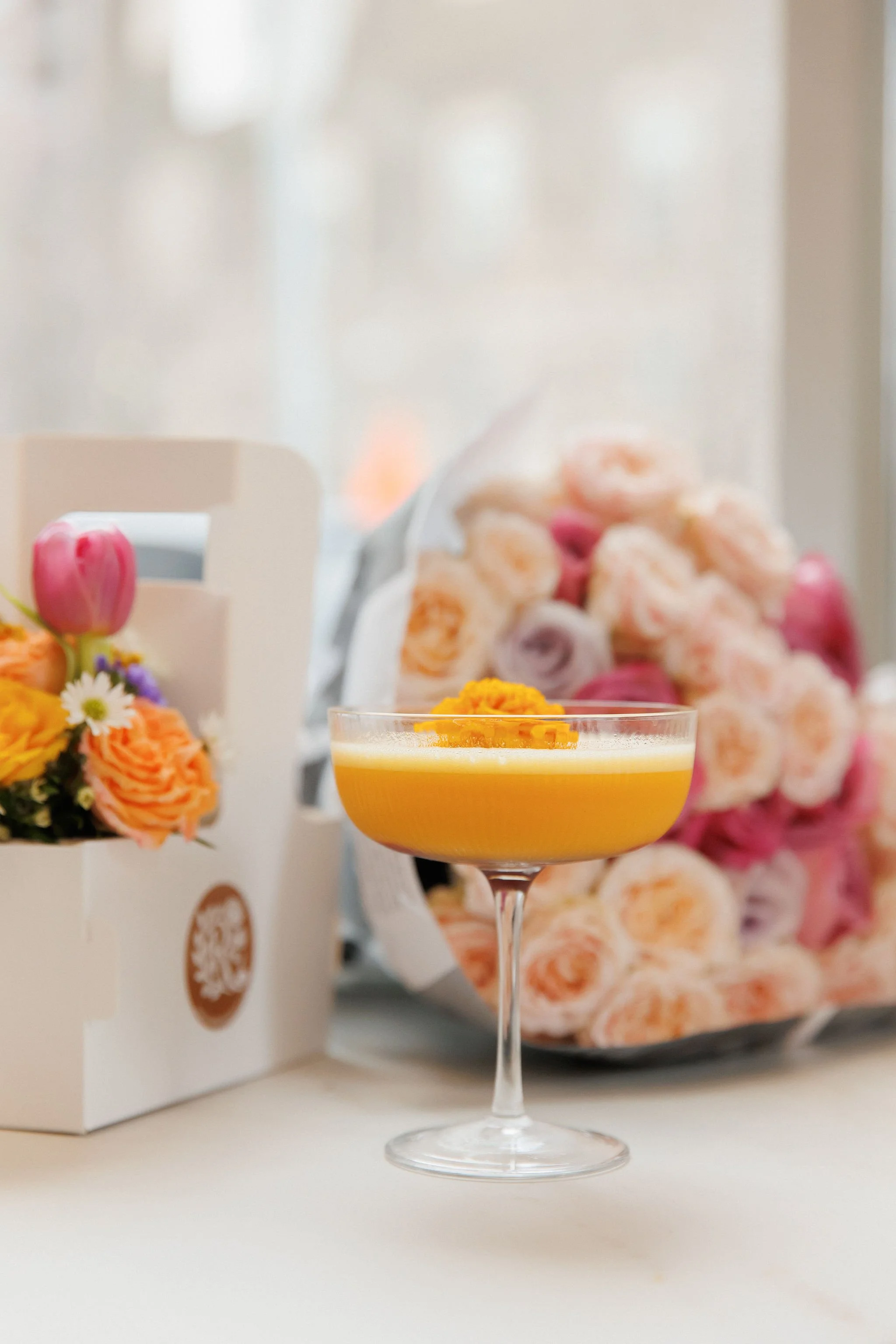 A cocktail glass with a yellow-orange drink topped with a small flower, set on a table next to a white floral gift box and a bouquet of pink, peach, and purple flowers.