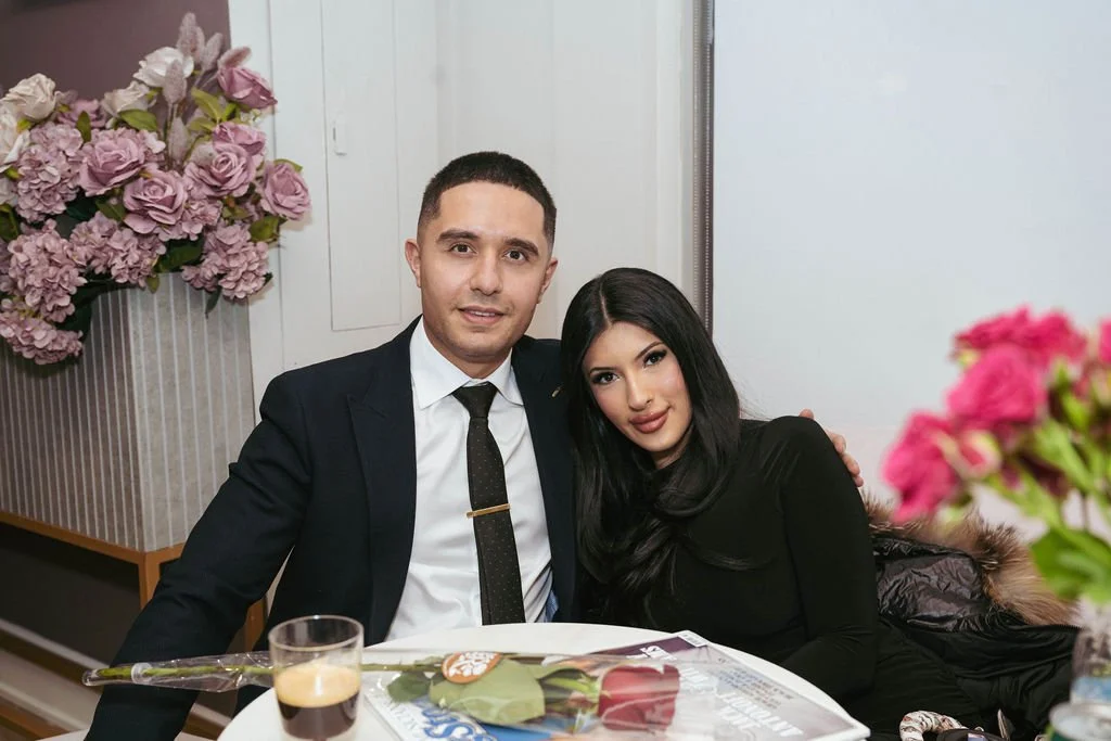 A man and a woman sitting together at a table, smiling. The man is wearing a dark suit with a white shirt and a black tie. The woman has long black hair and is dressed in black. There are pink flowers in a vase on the table, with a magazine and a gla