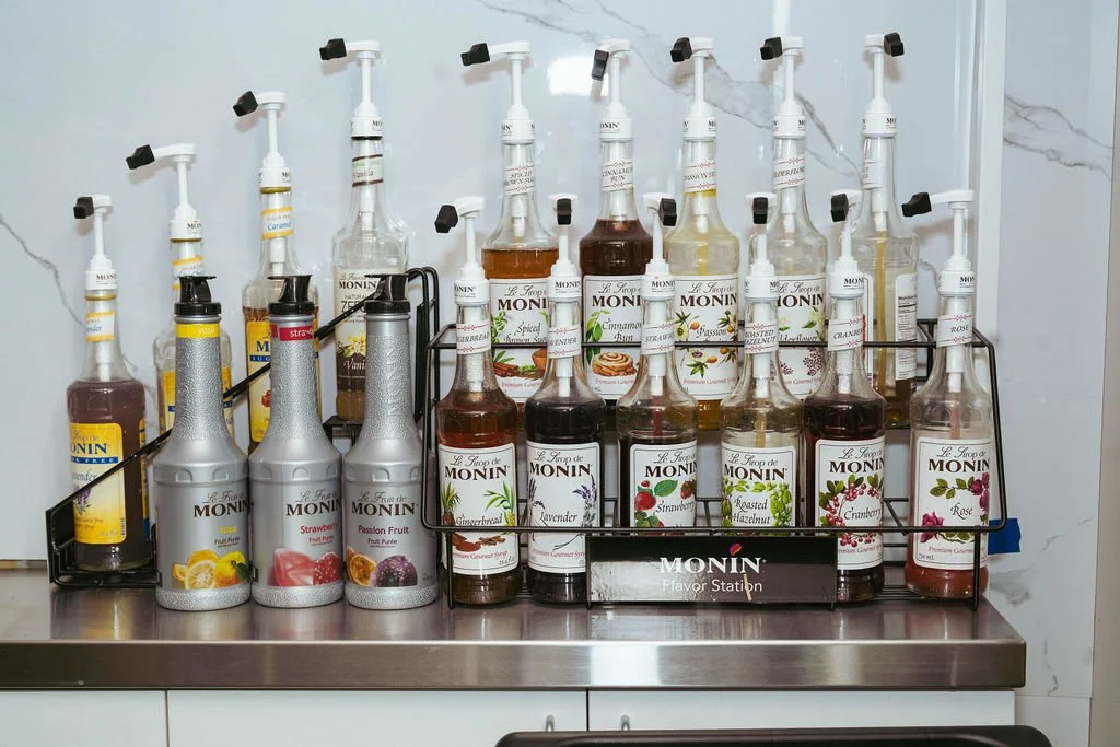 A collection of Monin flavored syrup bottles on a metal tray at a flavor station, including strawberry, passion fruit, gingerbread, lavender, strawberry, cinnamon, and raspberry flavors.