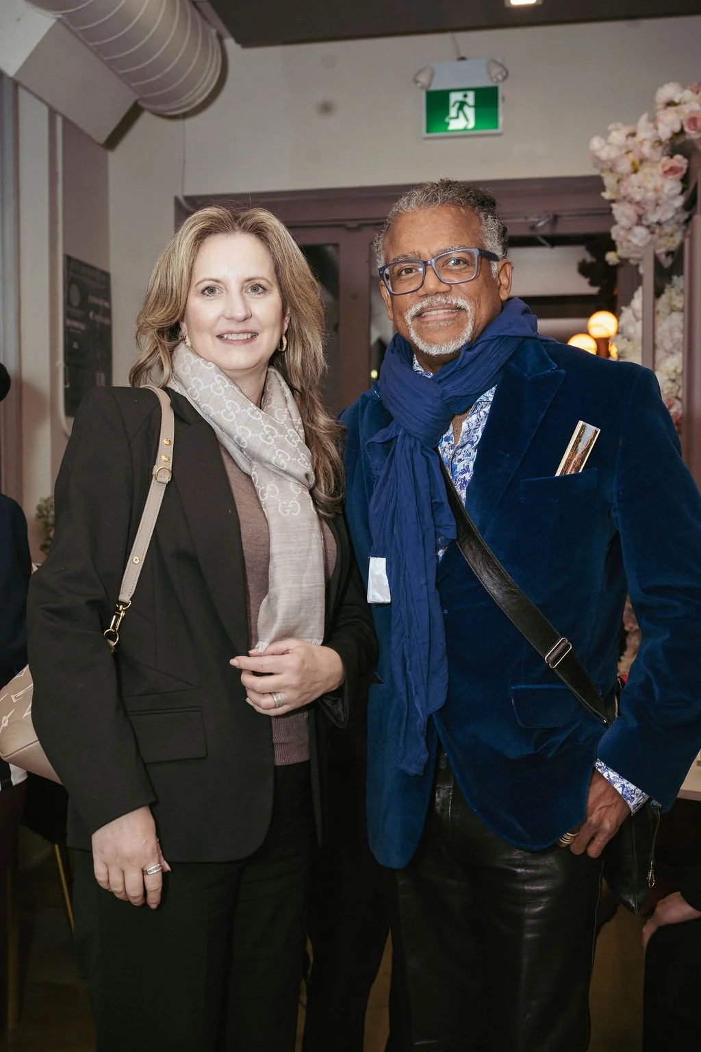 A woman and a man standing indoors, smiling at the camera. The woman has light brown hair, wearing a black blazer, beige scarf, and a ring on her left hand. The man has gray hair, glasses, a gray beard, wearing a blue velvet blazer, a patterned shirt