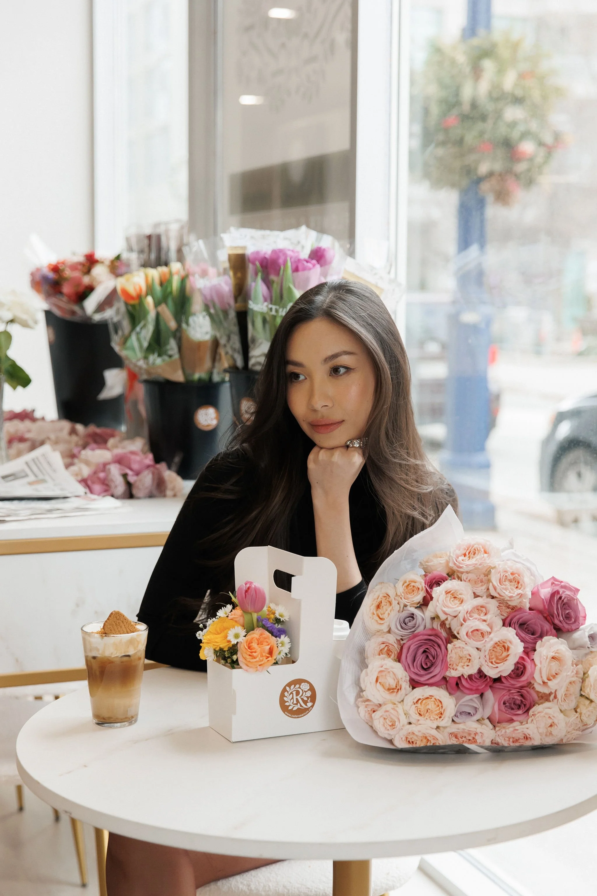 A woman sitting at a table with a bouquet of pink and peach roses, a box of flowers, and a glass of iced coffee inside a flower shop, with flowers and bouquets on display in the background.