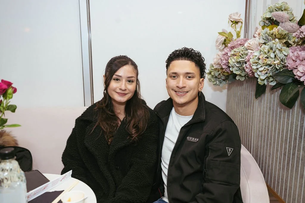 A young woman and man sitting together at a table in a restaurant, smiling at the camera, with pink and white flowers in the background.