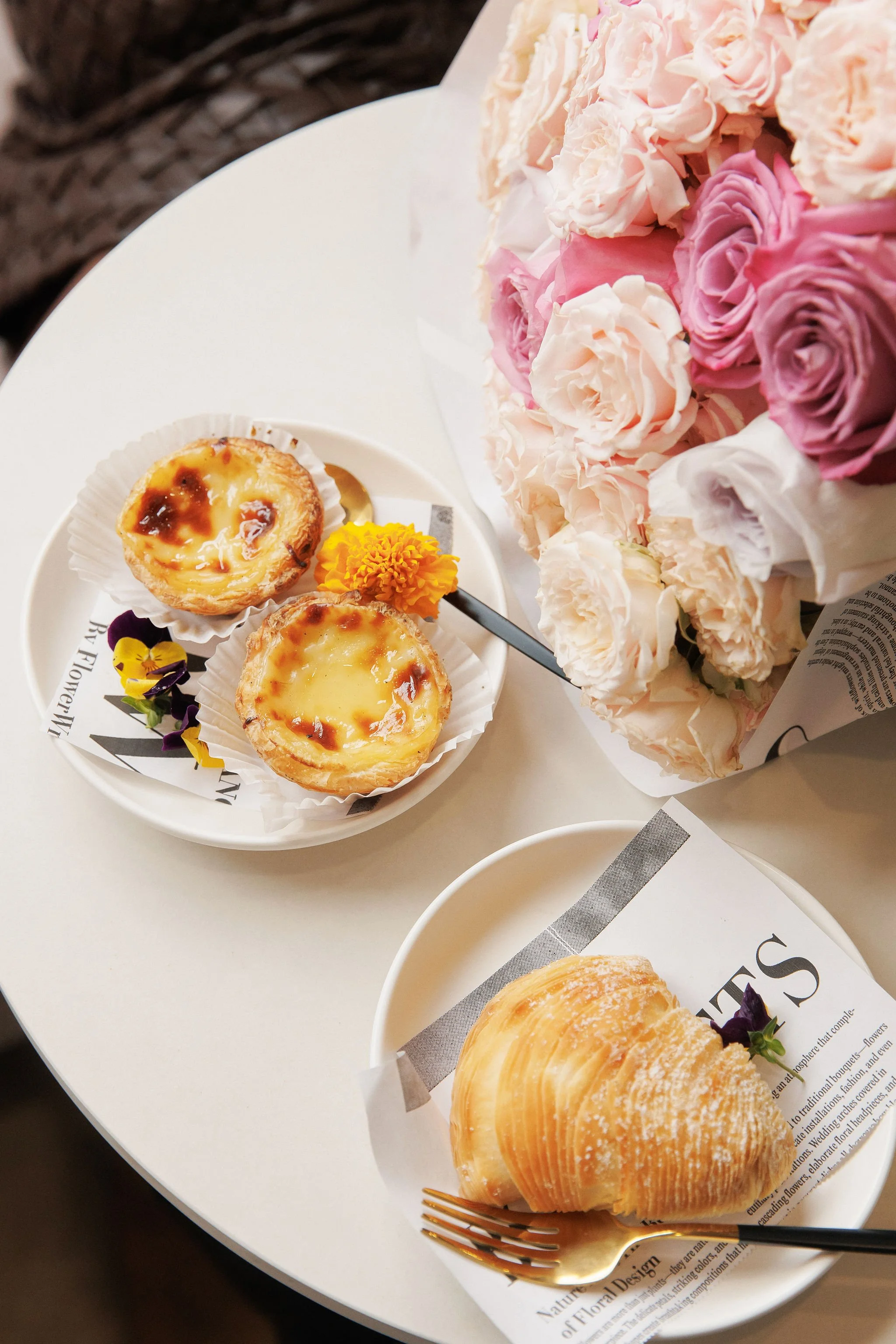 A white table with a bouquet of pink and cream roses, two plates with baked goods, and a fork. One plate has two egg tarts with a yellow flower and purple flower on the side, and the other has a croissant with a purple flower on the side.