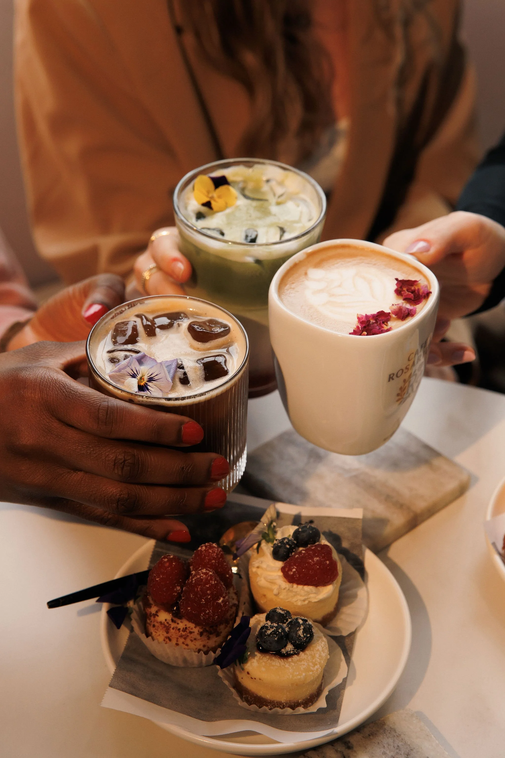 Three friends holding up glasses of iced coffee or tea, with a plate of assorted small desserts including cupcakes and fruit-topped tarts on a table.