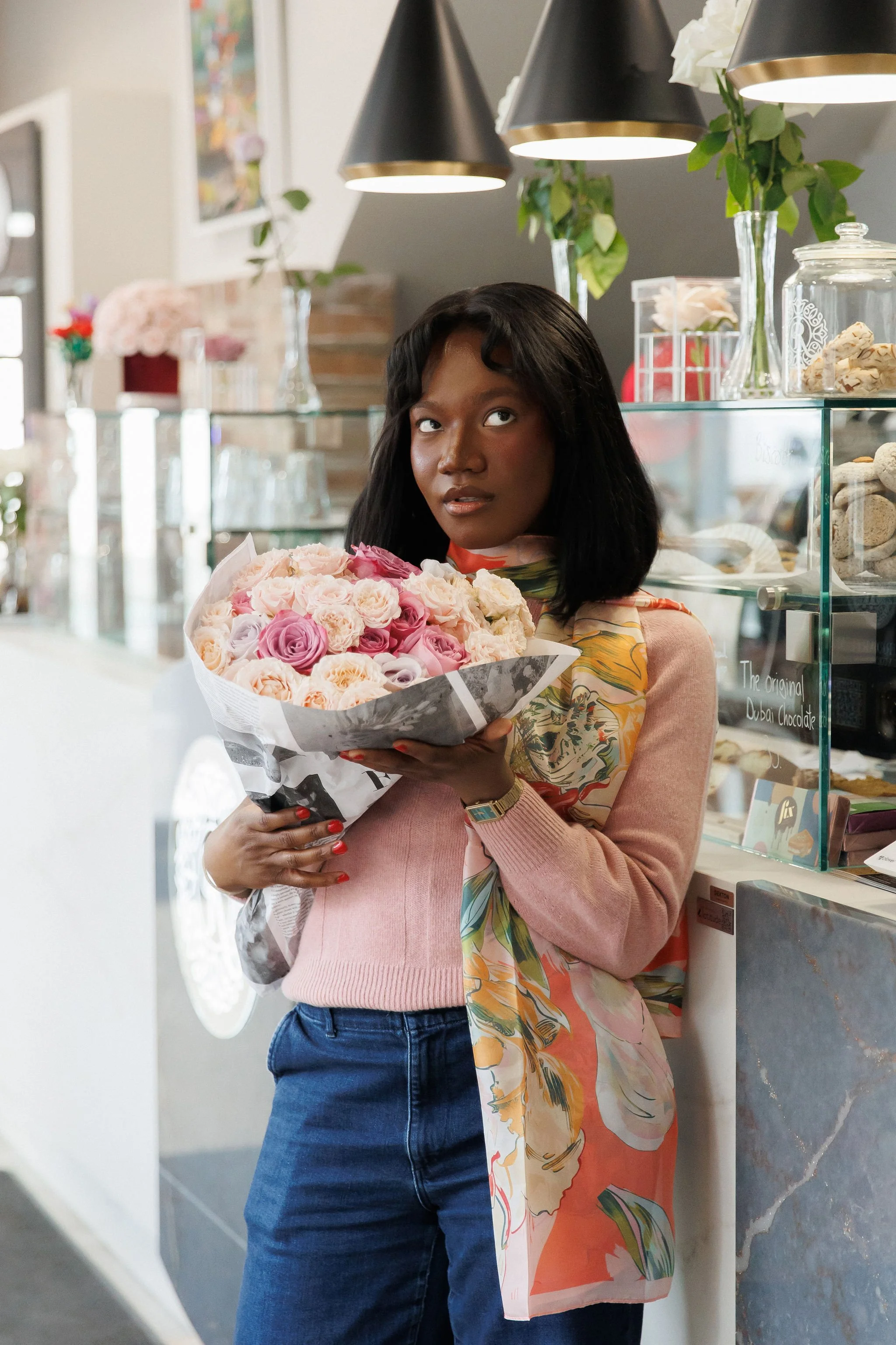 A woman holding a bouquet of pink and cream roses inside a flower shop or café.