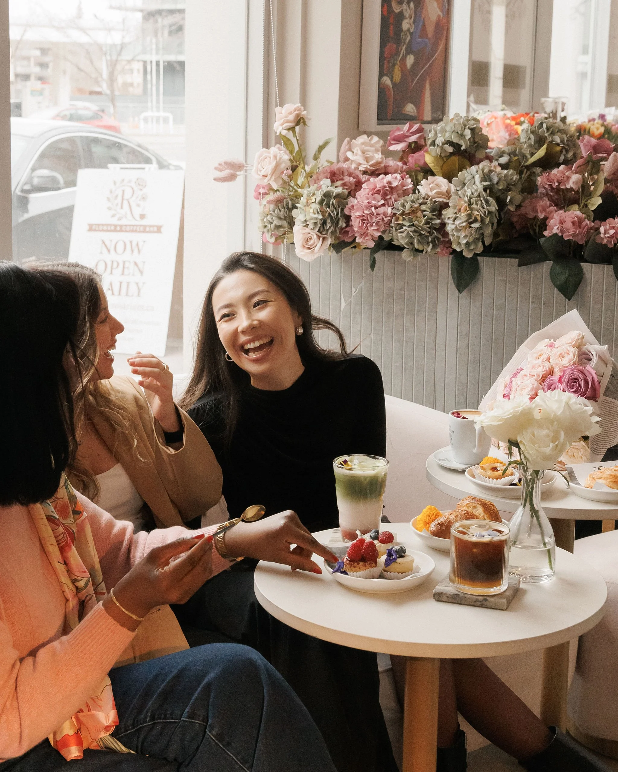 Three women having a cheerful conversation and enjoying desserts and drinks in a cafe decorated with pink and white flowers.