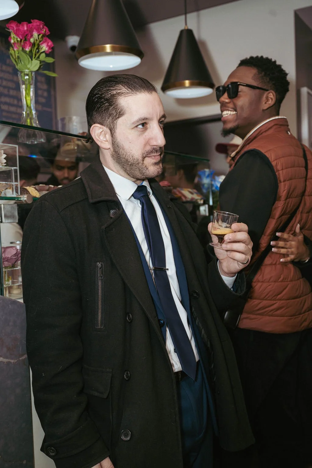 Two men at a social gathering, one holding a drink, in a modern indoor venue with pendant lighting and a flower arrangement in the background.