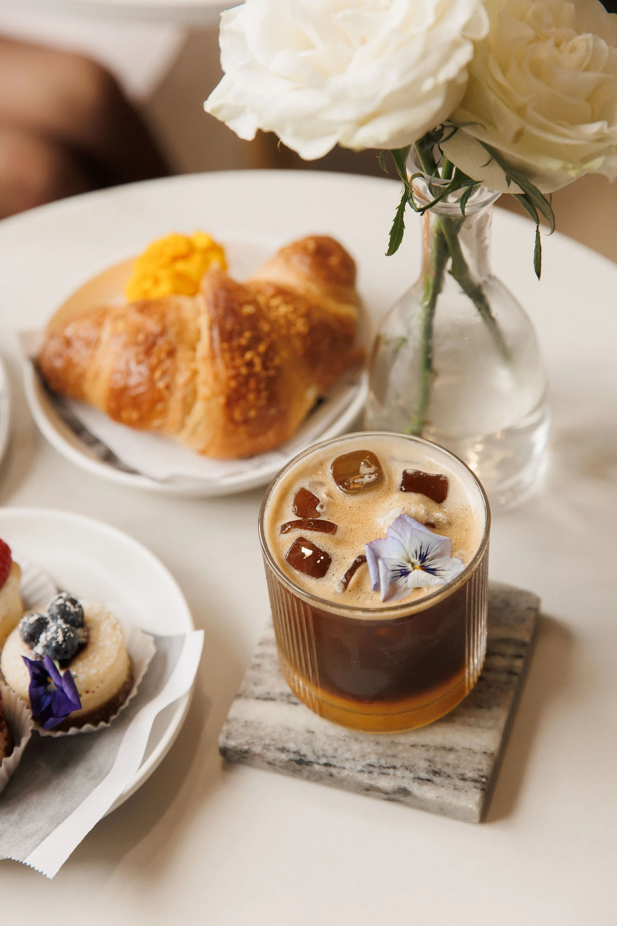 A glass of iced coffee with ice cubes and a purple flower garnish sits on a small marble coaster. Behind it, a white plate holds a croissant with a dollop of butter or honey. To the left, part of a dessert with berries and edible flowers is visible. 