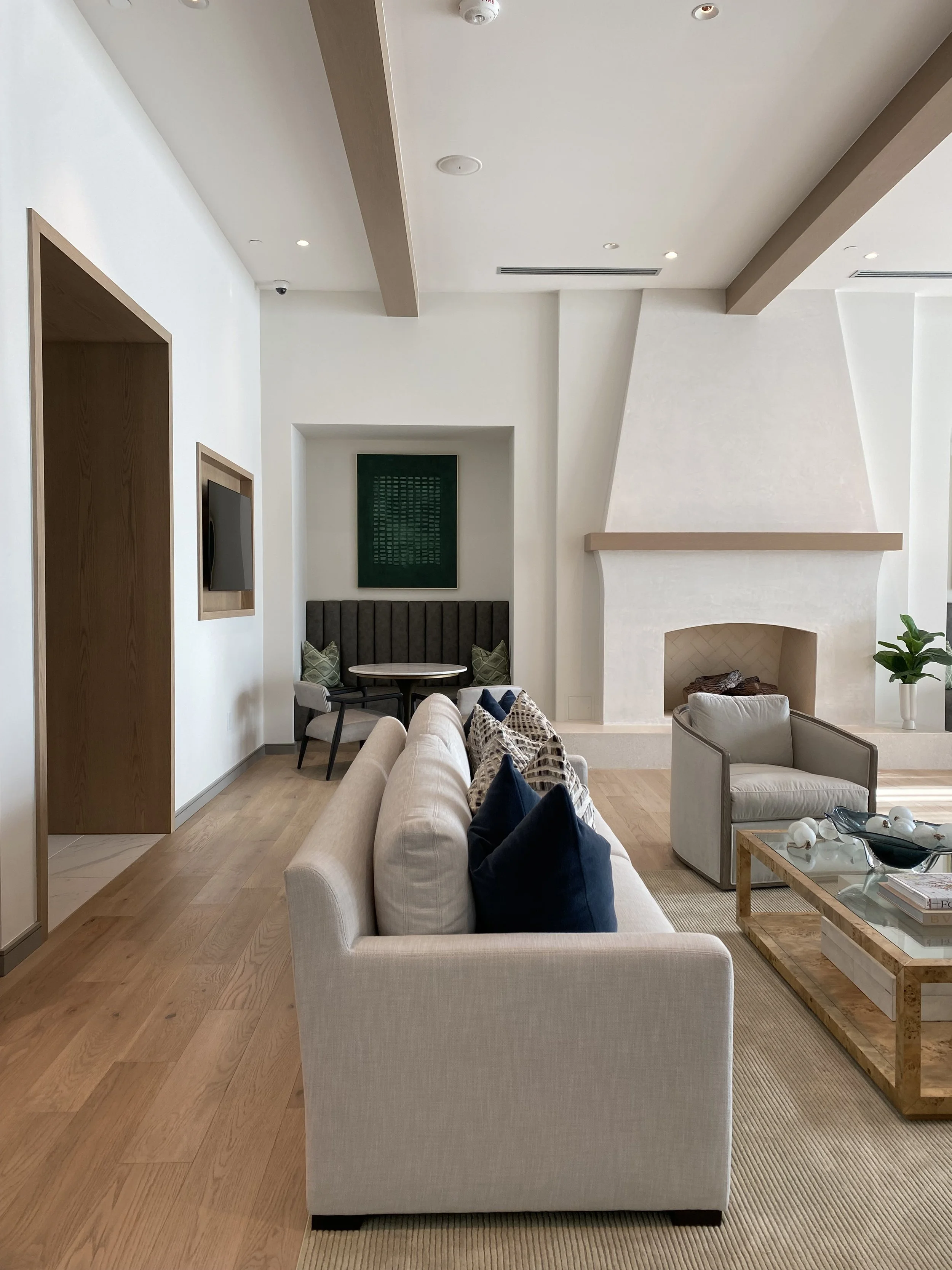 Modern living room with a light gray sofa, navy and patterned throw pillows, a glass coffee table with decor, an armchair, a large fireplace with a wooden mantle, and a small dining area with a table and chairs in the background. The room features wooden beams on the ceiling and hardwood floors.