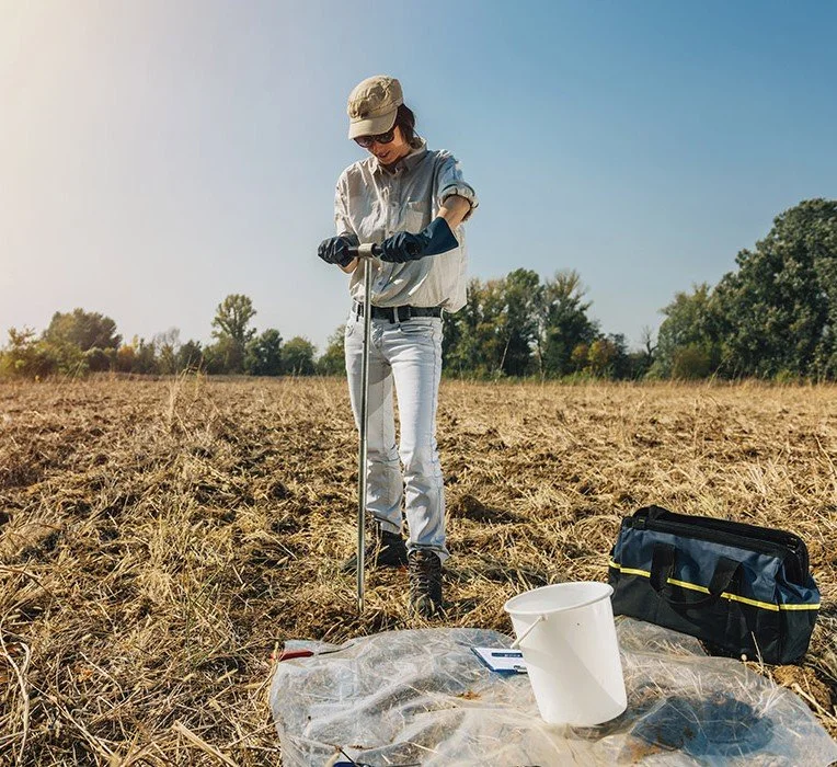 Person conducting soil sampling in a field with equipment and bucket.