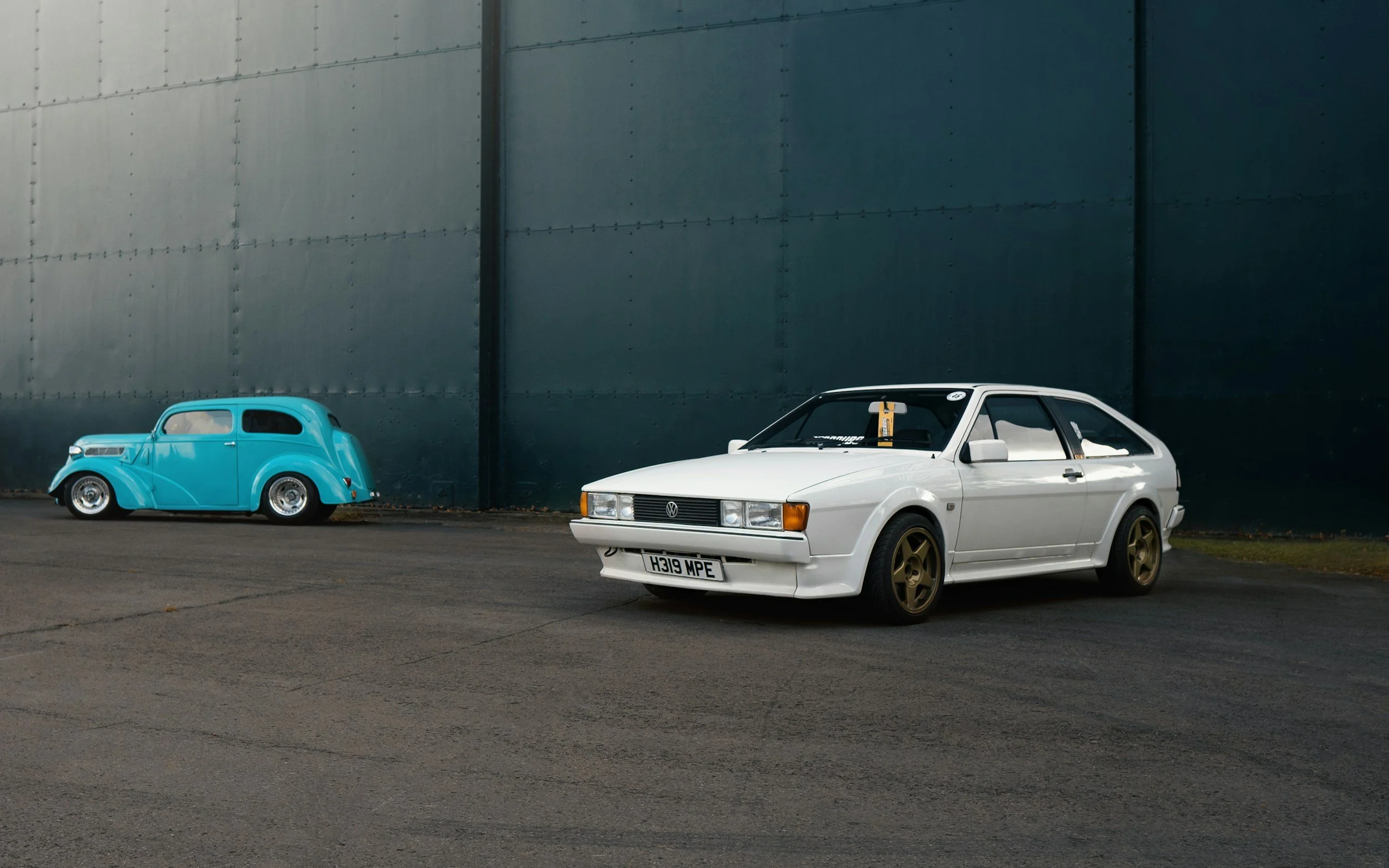A vintage white coupe car with gold wheels parked on a paved surface, with a rusty blue vintage car with a rounded body shape parked nearby against a blue industrial wall.