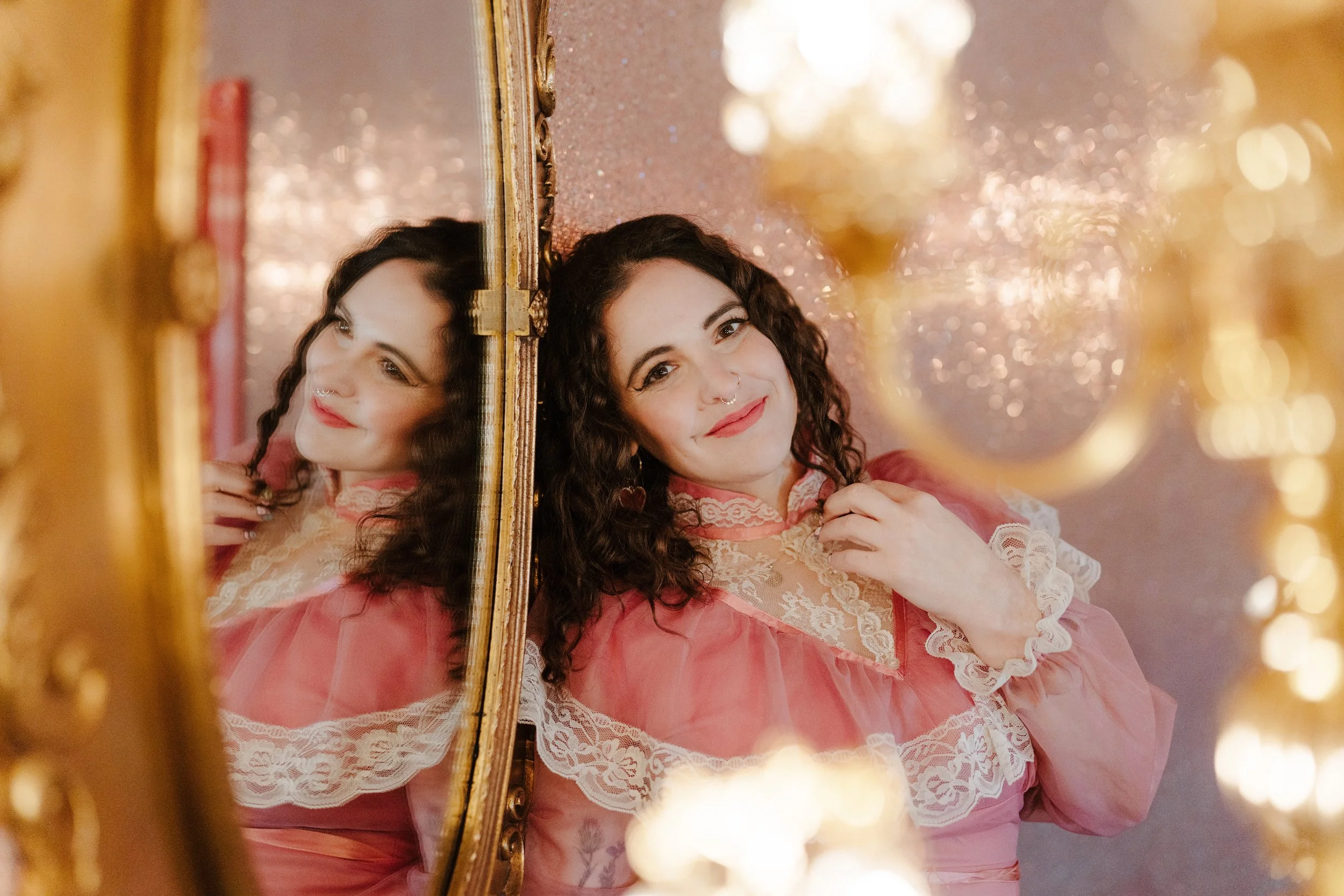 Photo of Ilana, a white woman with brown curly hair. she is leaning against a wall and smiling at the camera. In the foreground is a gold chandelier, and you can see her reflection in a gold framed mirror. Her dress is pink and high necked with lace.