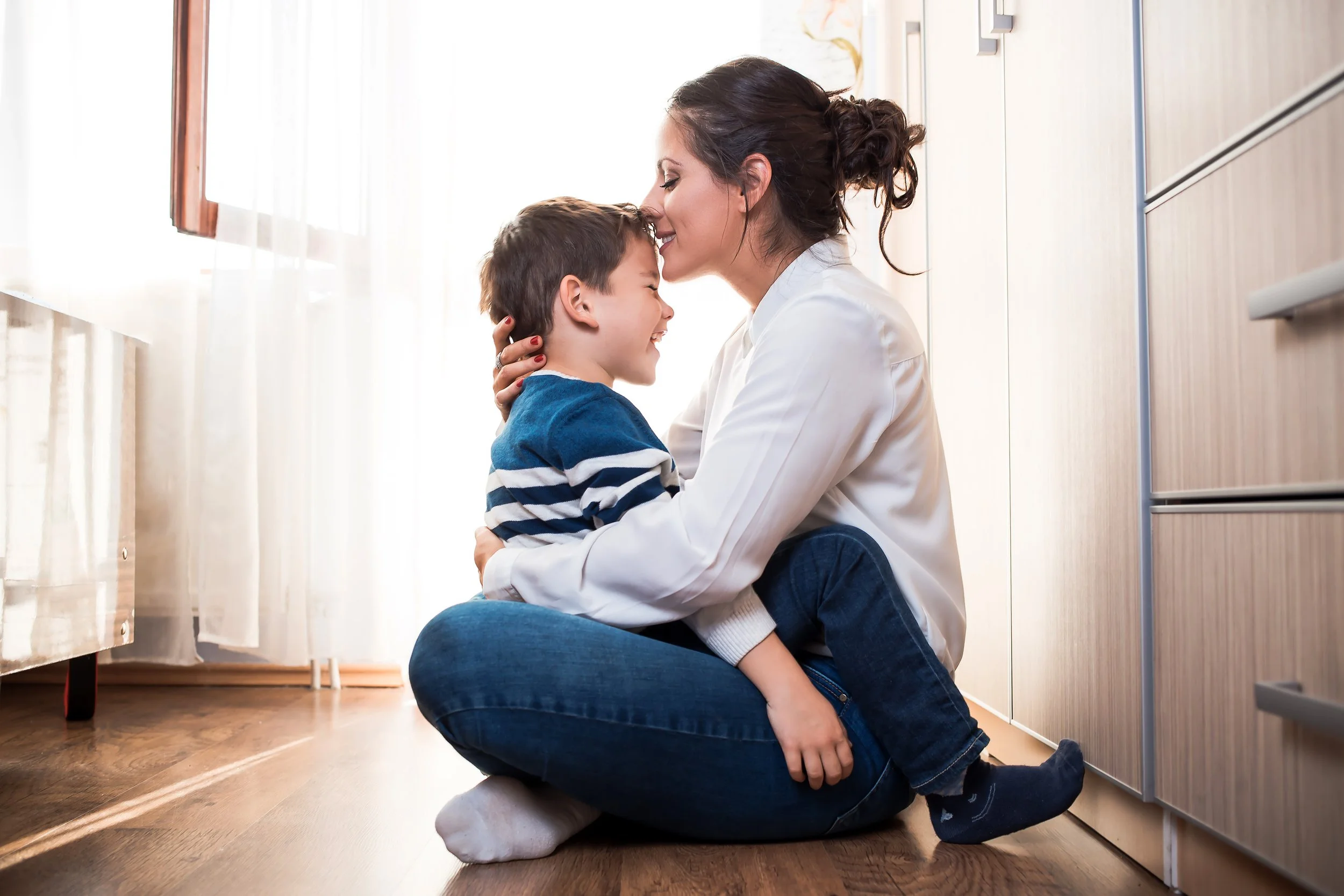 A woman and a young boy share a tender moment in a kitchen, with the woman kneeling and gently kissing the boy's forehead as he smiles with his eyes closed.