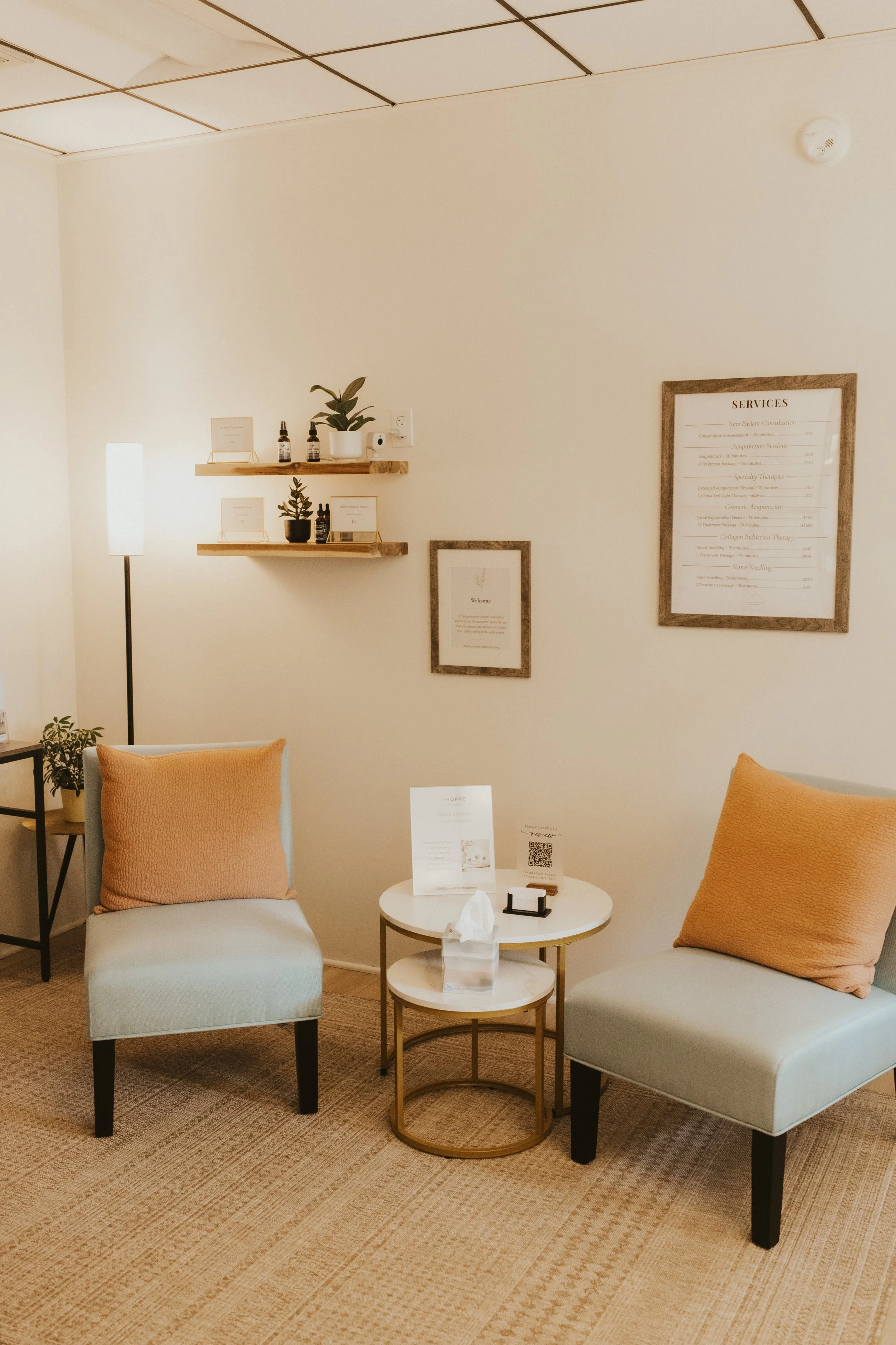 white door on left with bell on doorknob, and window on right with white blinds, two aqua chairs in front of window, with white and tan checkered pillows, and a white desk with gold accents in front of chairs