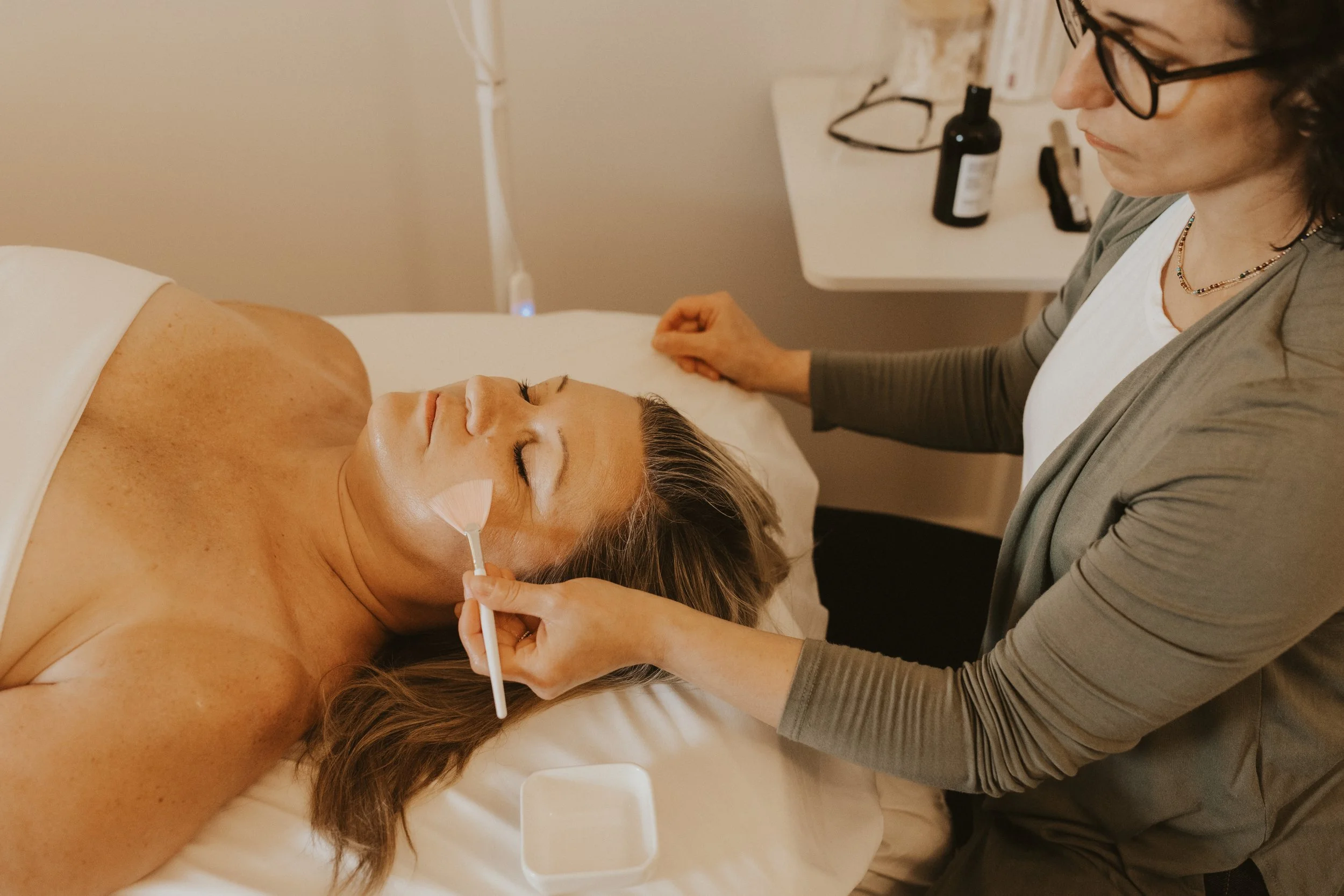 white woman lying on treatment table with white towel wrapped around her chest. white towel rolled up under her head, candles in the background, and practitioner's hands resting on patient's head