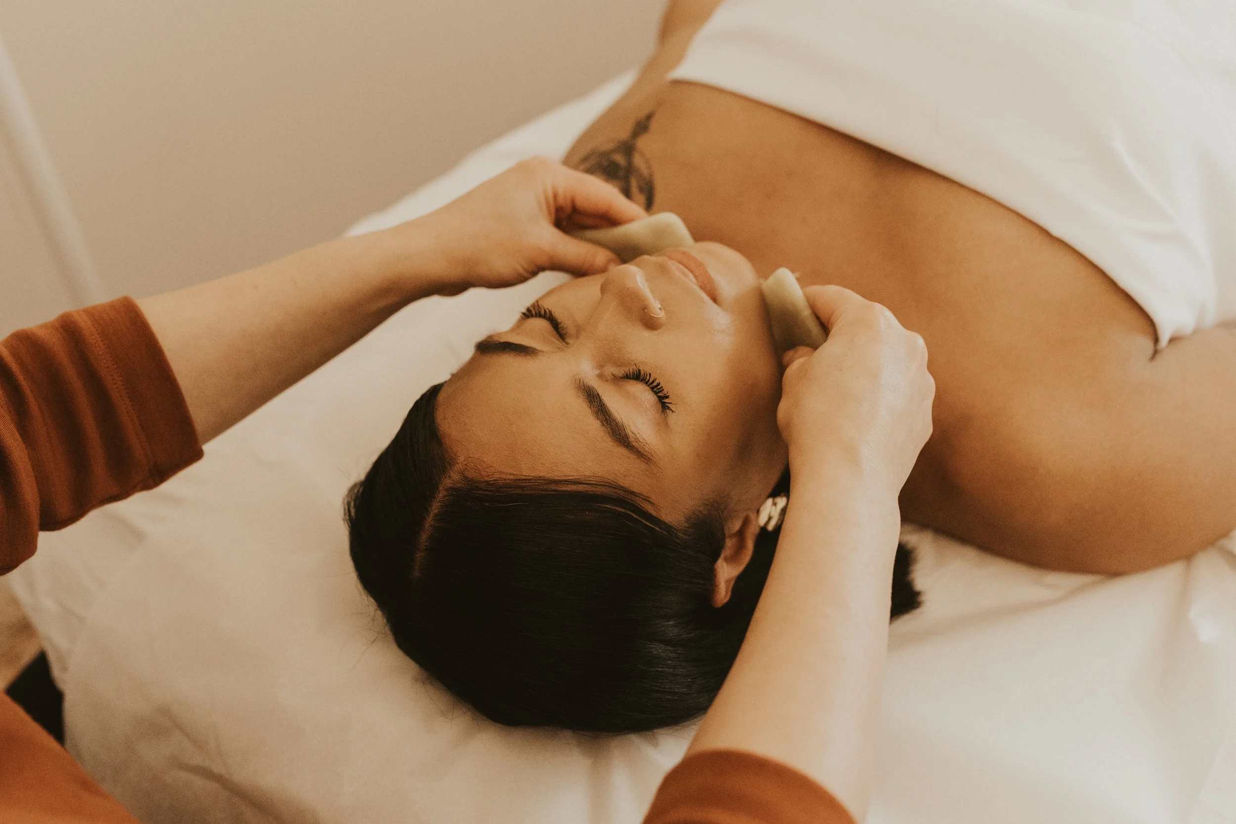 older white woman patient resting on white treatment table, wrapped in white towel, and white practitioner with short brown hair and glasses, wearing denim shirt, performing facial cupping on patient