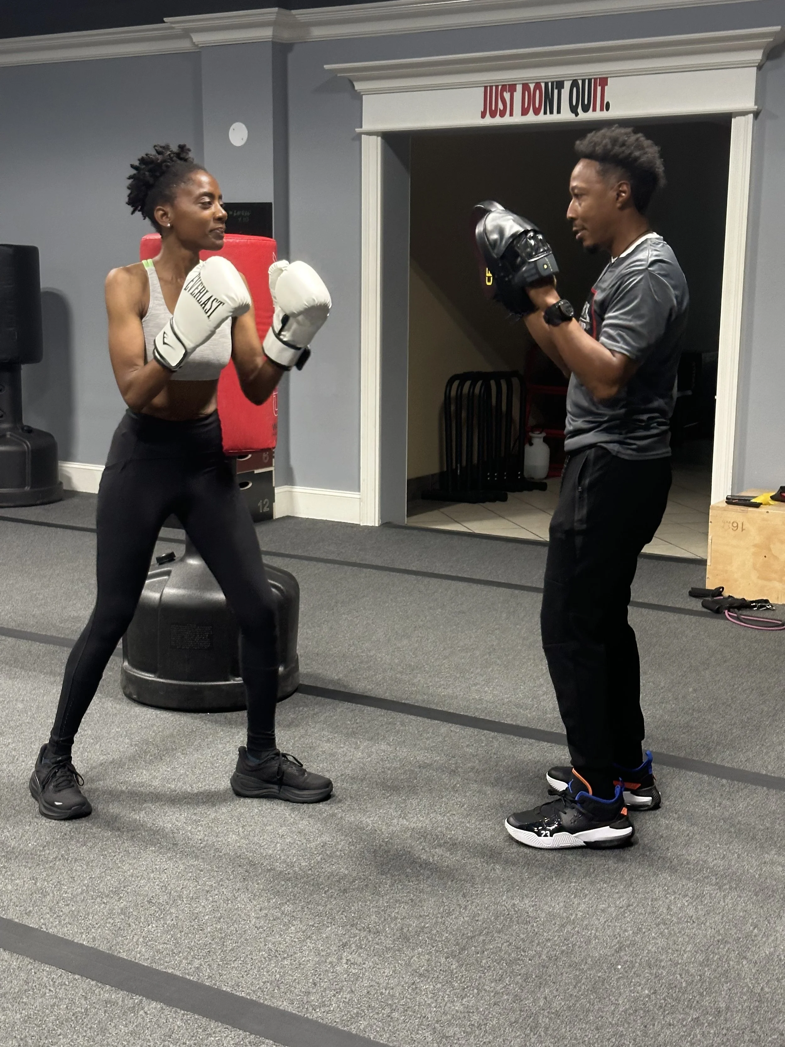 Two people are practicing boxing in a gym. The woman on the left is wearing boxing gloves and workout clothes, facing the man on the right, who is wearing a t-shirt, pants, and holding boxing mitts. They are standing on a gray workout mat with a moti