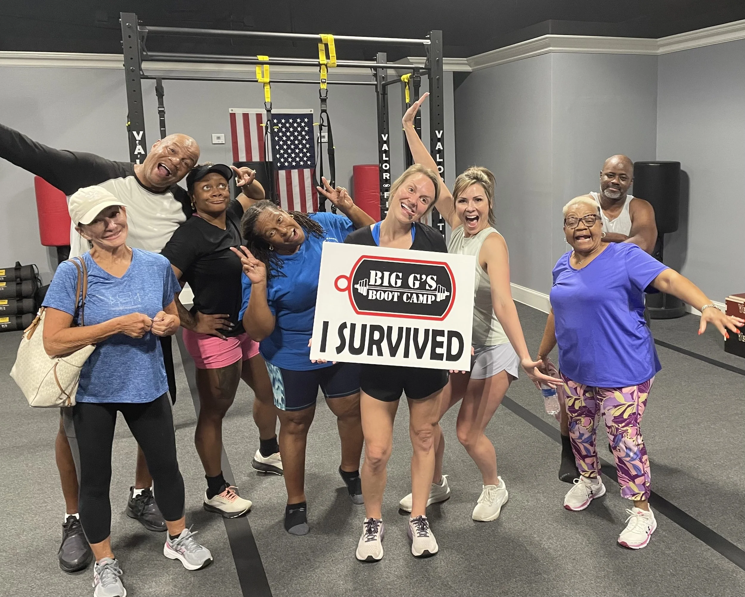 Group of diverse people celebrating after boot camp, holding a sign that says 'Big G's Boot Camp I Survived' in a gym with fitness equipment.