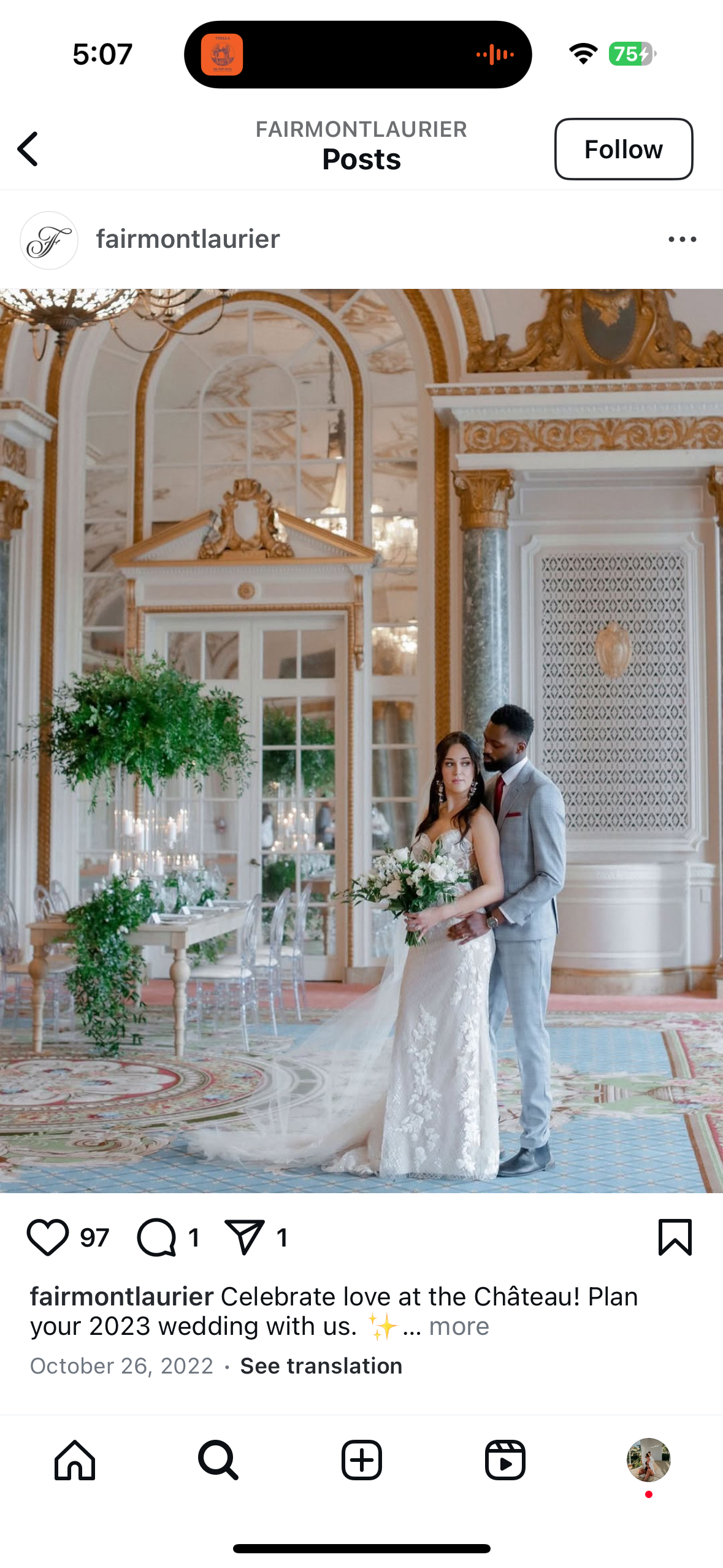 A bride and groom pose elegantly in a luxurious, ornate room with high ceilings and intricate decor. The bride wears a white lace gown and holds a bouquet, while the groom is in a gray suit with a red tie. The setting features chandeliers, lush greenery, and a decorative floor.