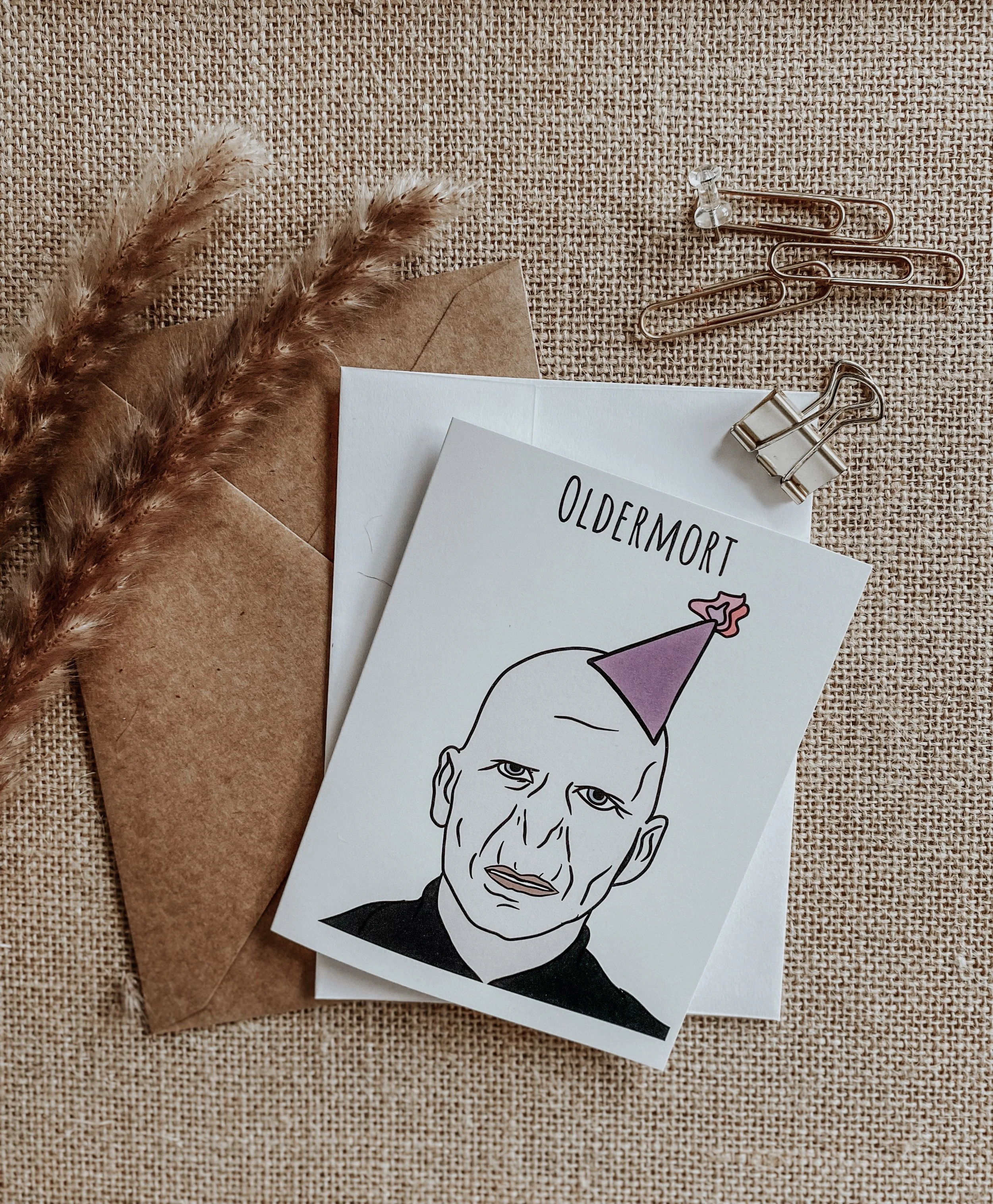 Birthday card with "Oldermort" illustration featuring a bald character with a party hat, on a burlap surface with paper clips, dried grass, and envelopes.