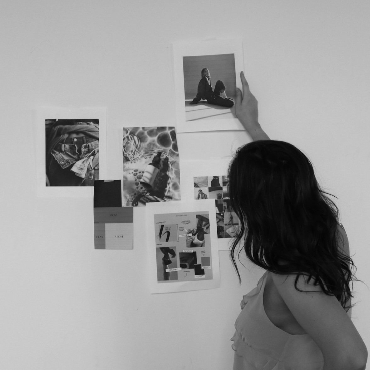 A person with long hair looking at a wall with various black and white pictures and photos attached, possibly arranging them.