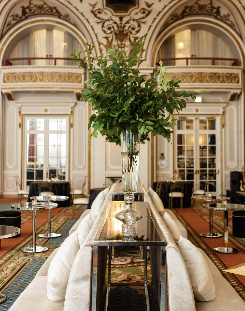 Luxurious hotel lobby with ornate ceiling, large vase of green leaves on a mirrored table, and elegant seating.