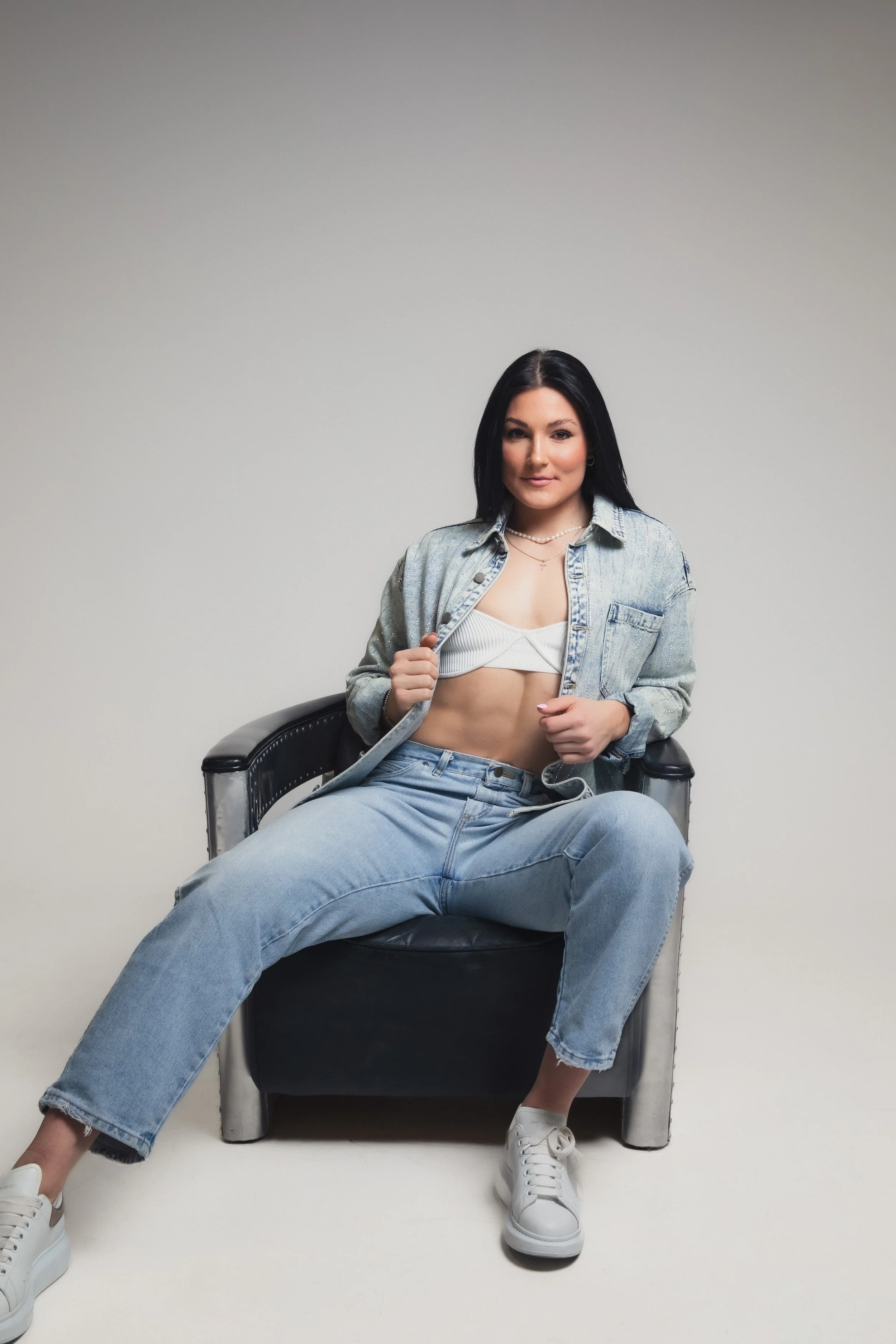 Person sitting in a chair wearing a denim outfit and white sneakers against a neutral background.