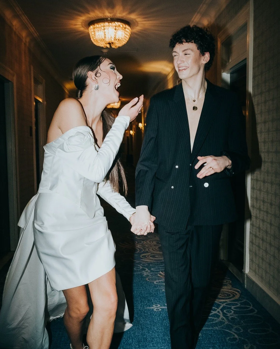 A woman and a man dressed in formal attire walking and laughing in a dimly lit hallway with chandeliers.