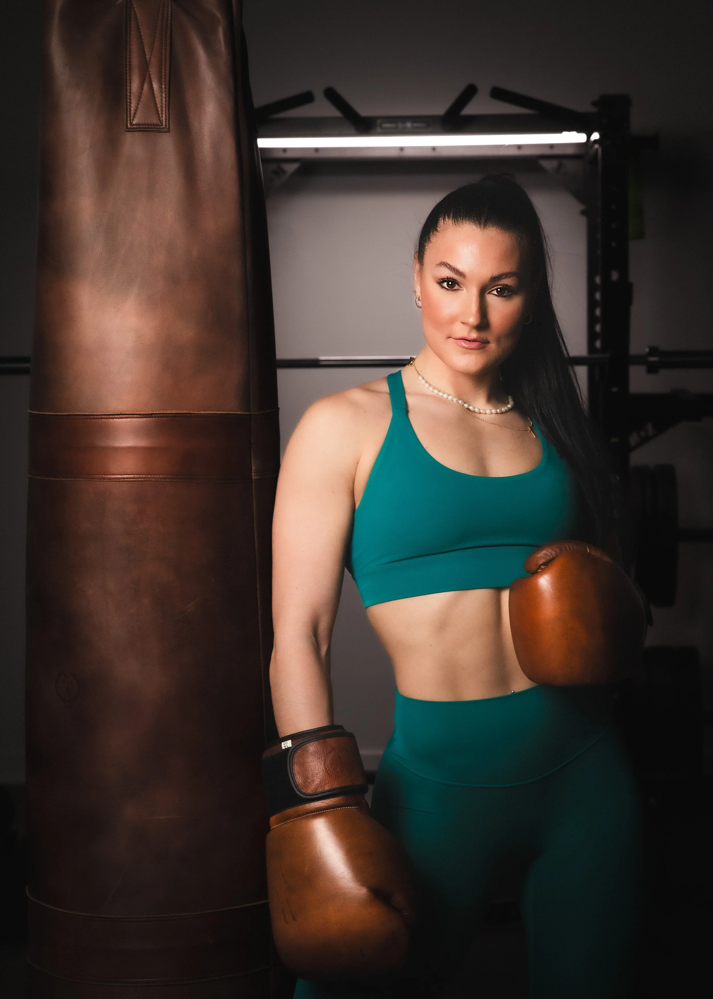 Woman in teal sports attire and brown boxing gloves standing next to a punching bag in a gym.