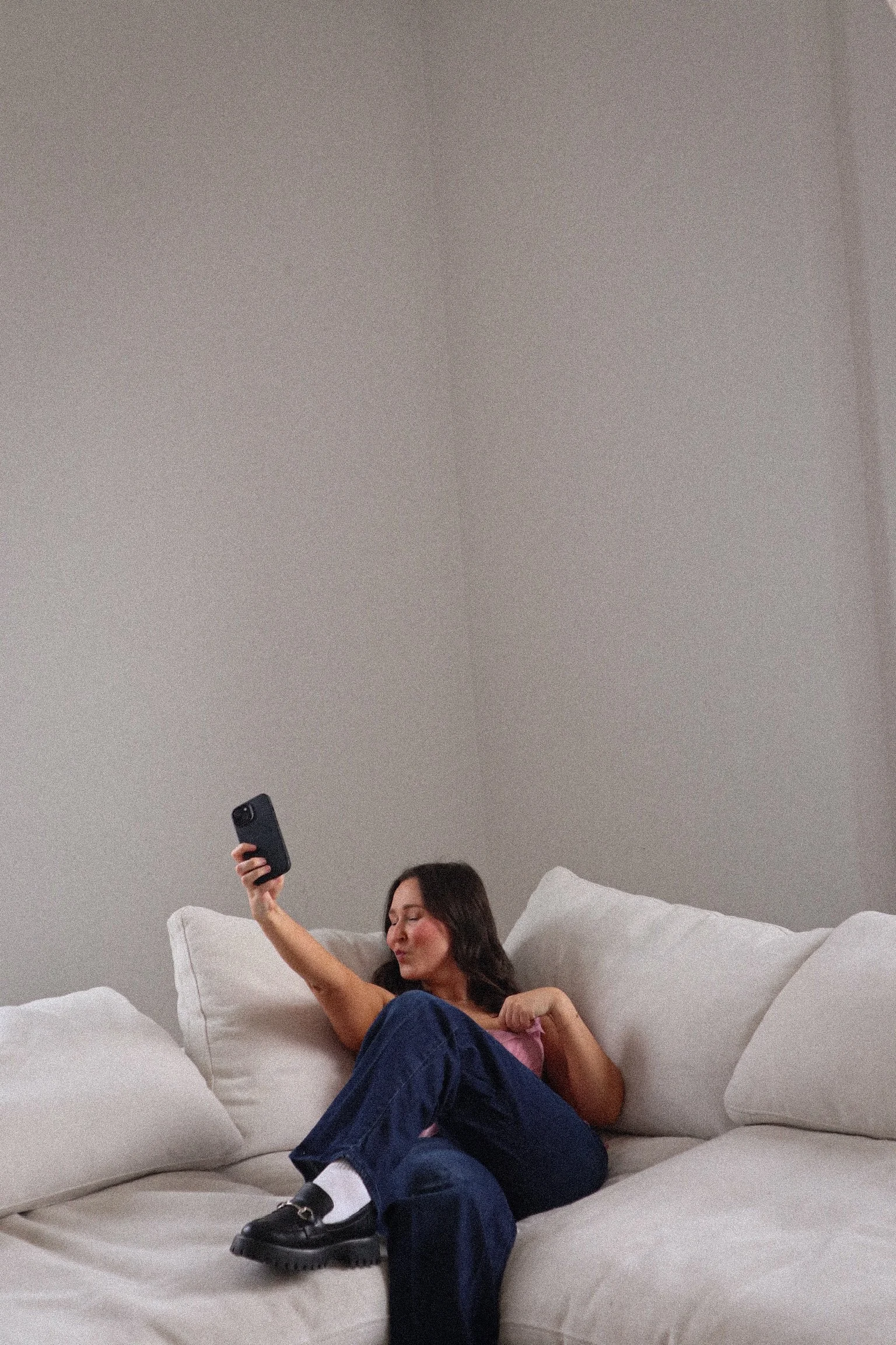 A person sitting on a white couch taking a selfie with a smartphone, wearing a sleeveless top and jeans.