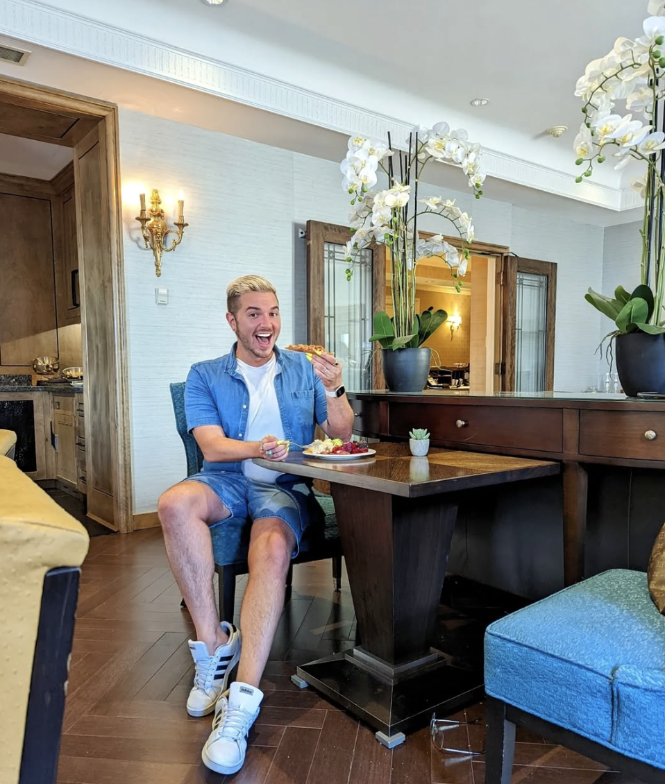 Person sitting in a room, eating pizza and holding a fork with a breakfast plate on the table.