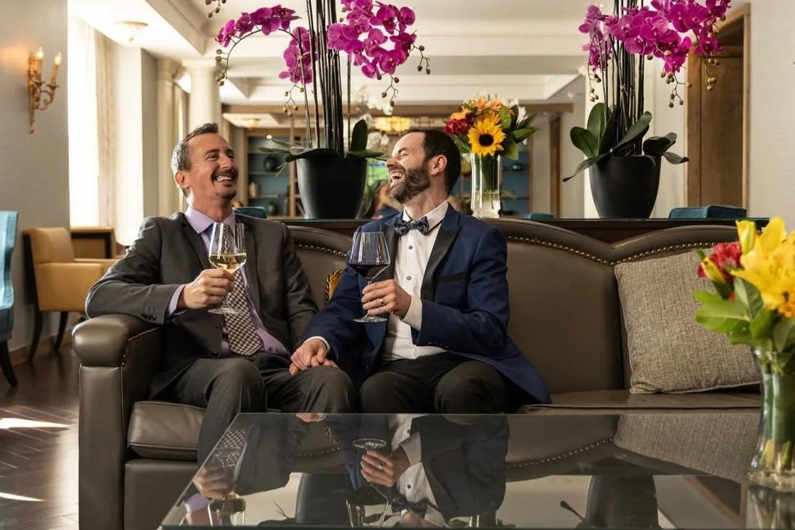 Two men in suits laughing and holding wine glasses, sitting on a couch in an elegant room decorated with flowers and orchids.