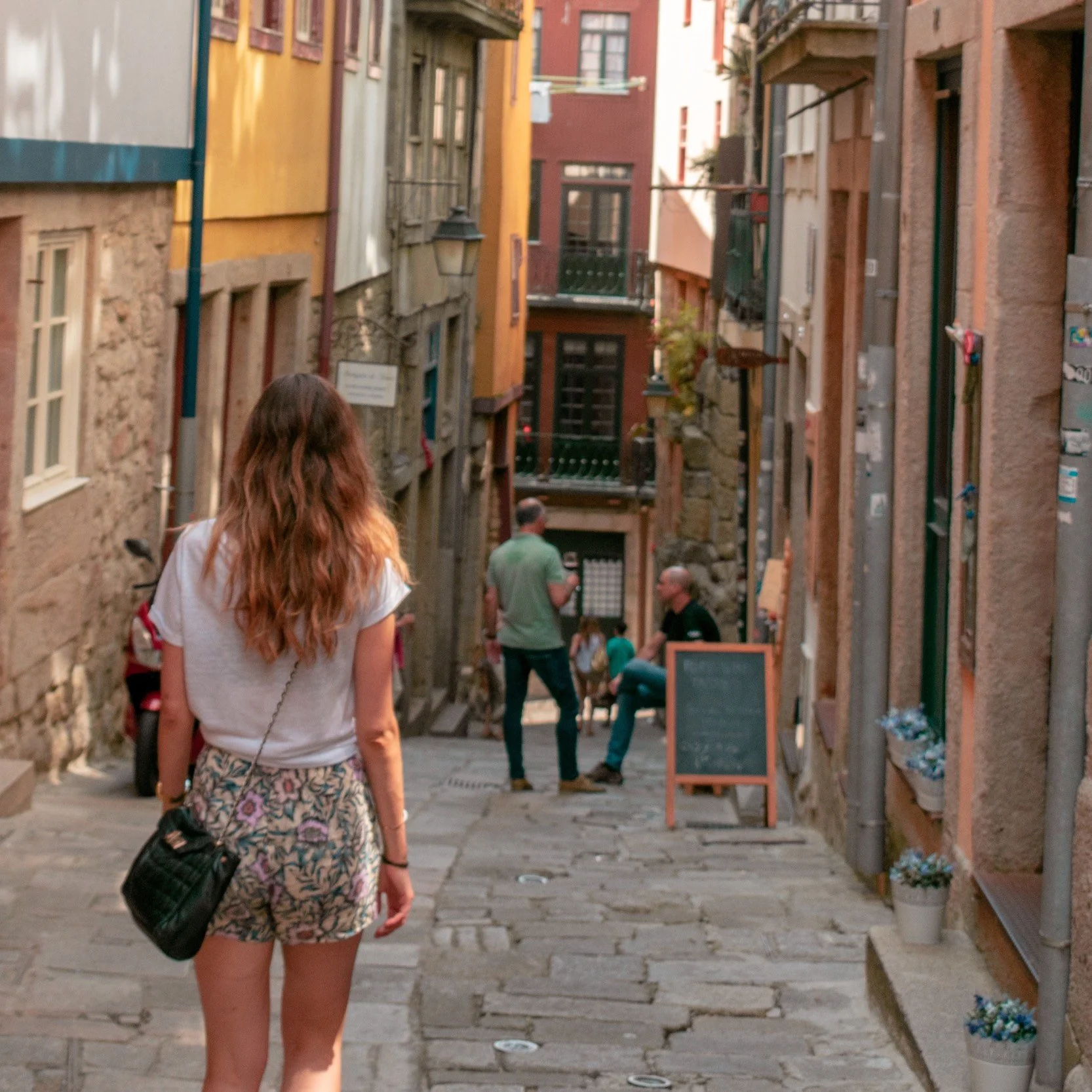 A woman with long, wavy brown hair wearing a white top and patterned shorts walks down a narrow, cobblestone street with colorful old buildings on either side. There are people in the distance, some standing and some sitting, with a small chalkboard sign on the sidewalk. The scene appears to be in a historic or European-style neighborhood.
