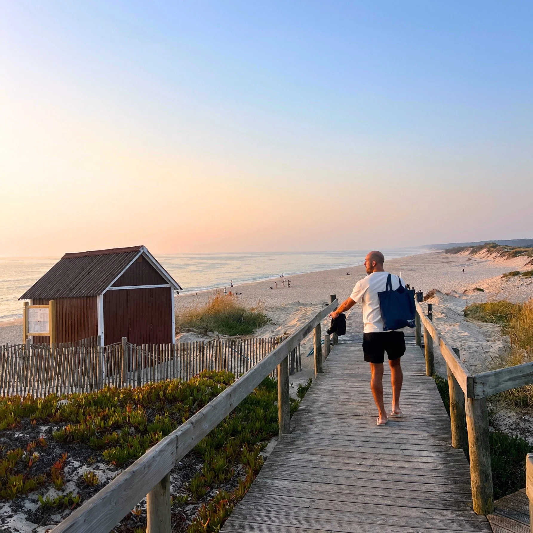 A man walking on a wooden boardwalk towards the beach at sunset, carrying a blue bag and a black hat, with a red beach hut nearby and sand dunes in the background.