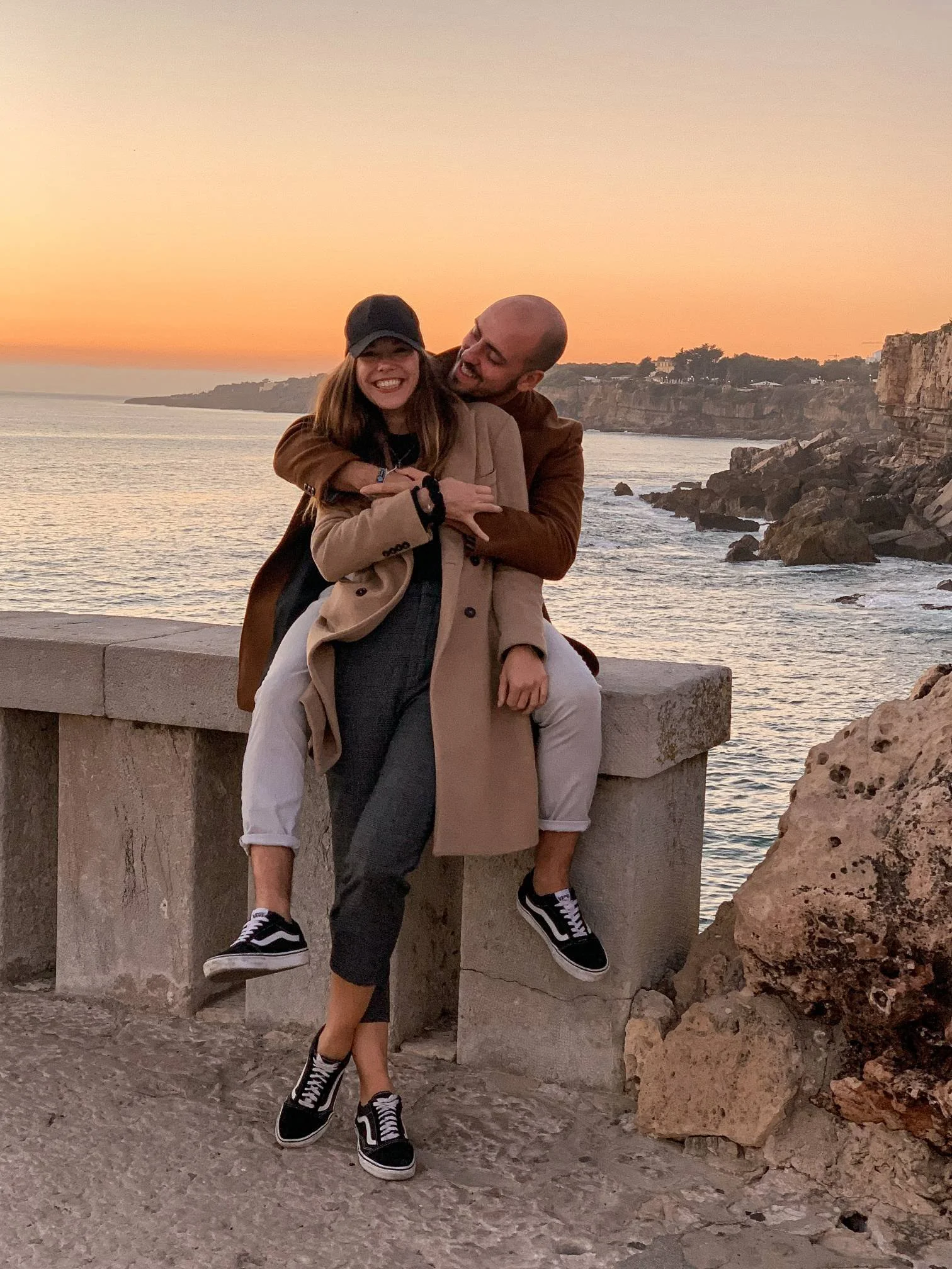 A smiling woman and a man hugging with a sunset over the ocean, rocky cliffs, and a stone bench in the background.