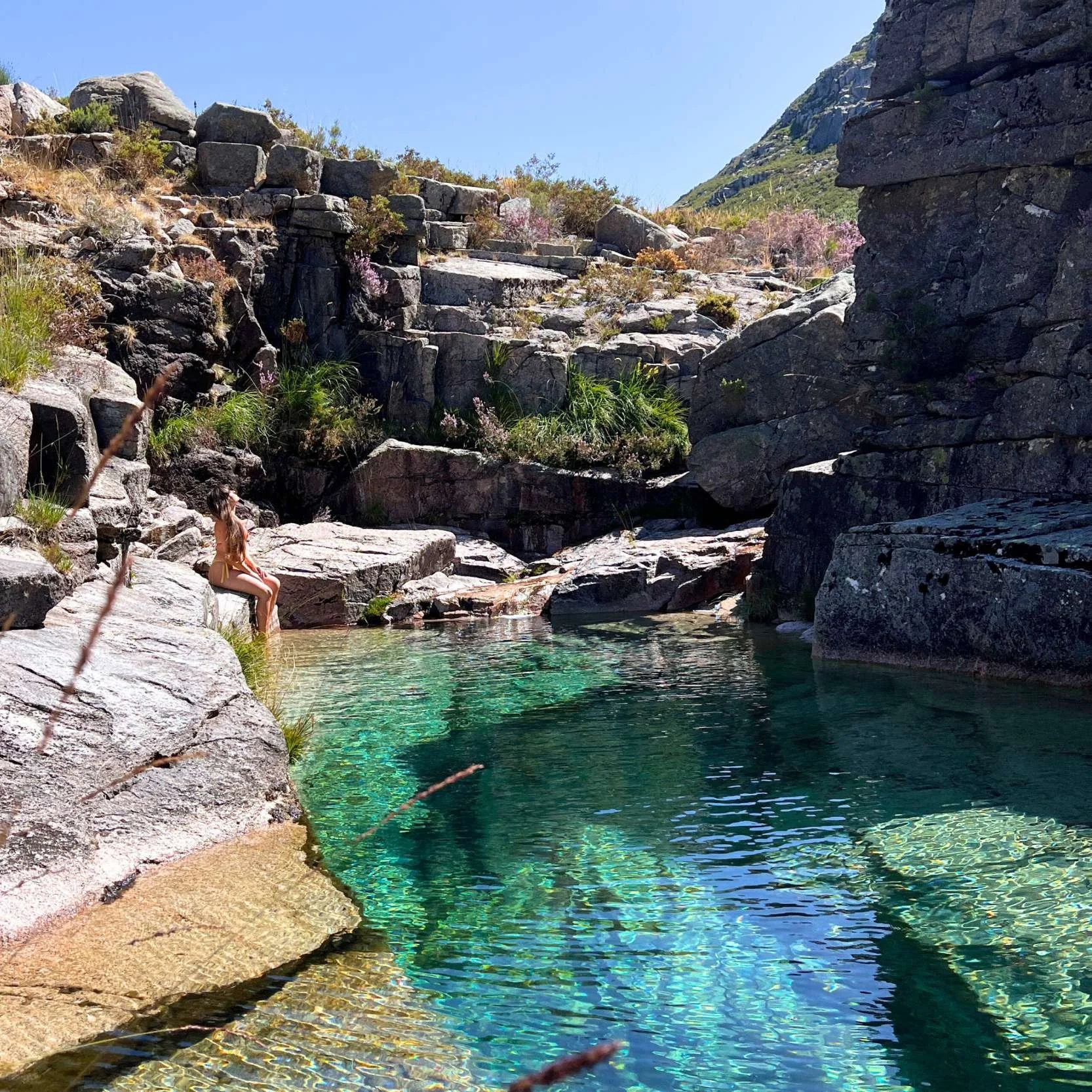 A girl sitting on rocks beside a clear, turquoise stream in a rocky canyon with plants and a mountain in the background.