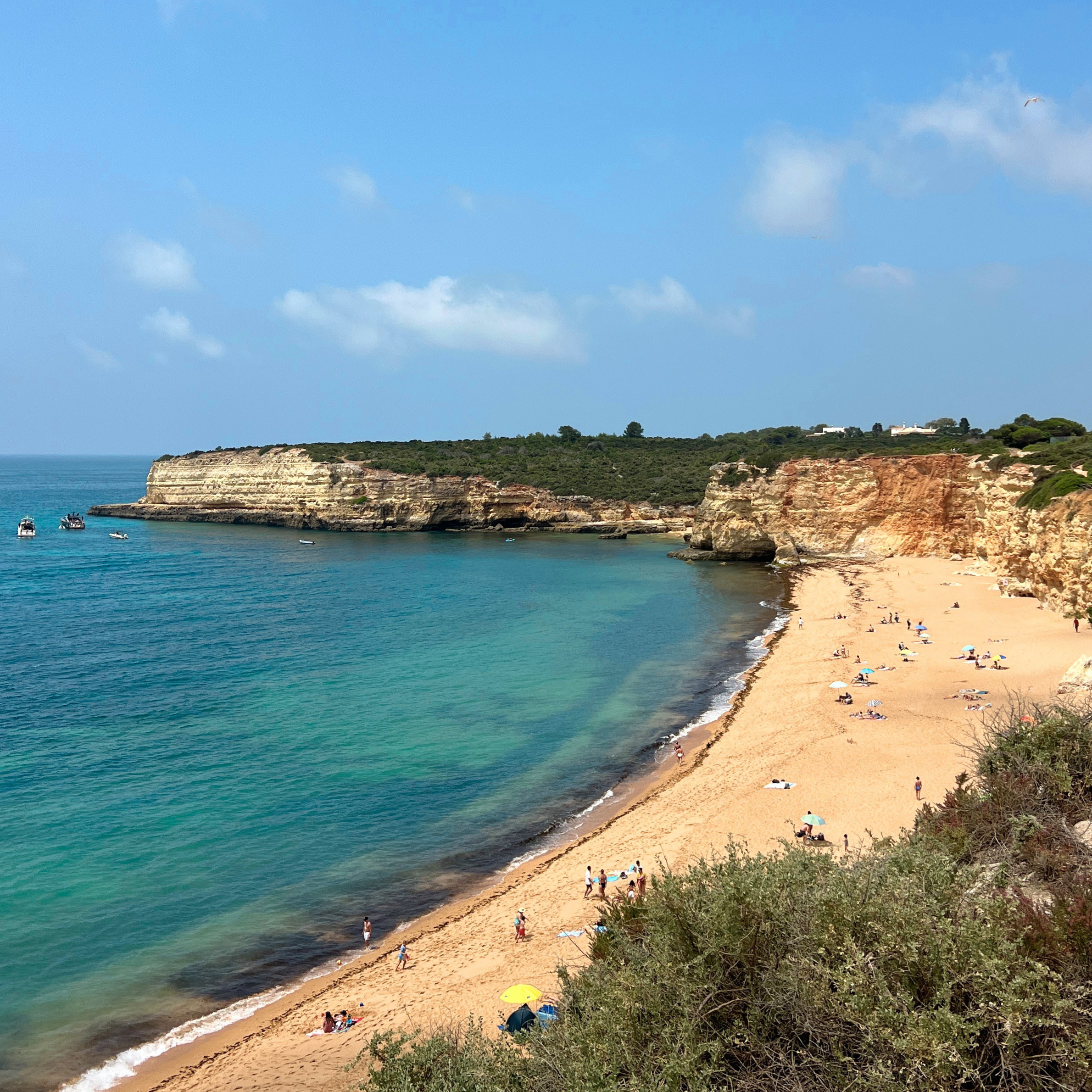 A beach with golden sand, turquoise water, and rocky cliffs overlooking the ocean. People are relaxing on the beach under umbrellas and walking along the shoreline. The sky is clear with a few clouds.