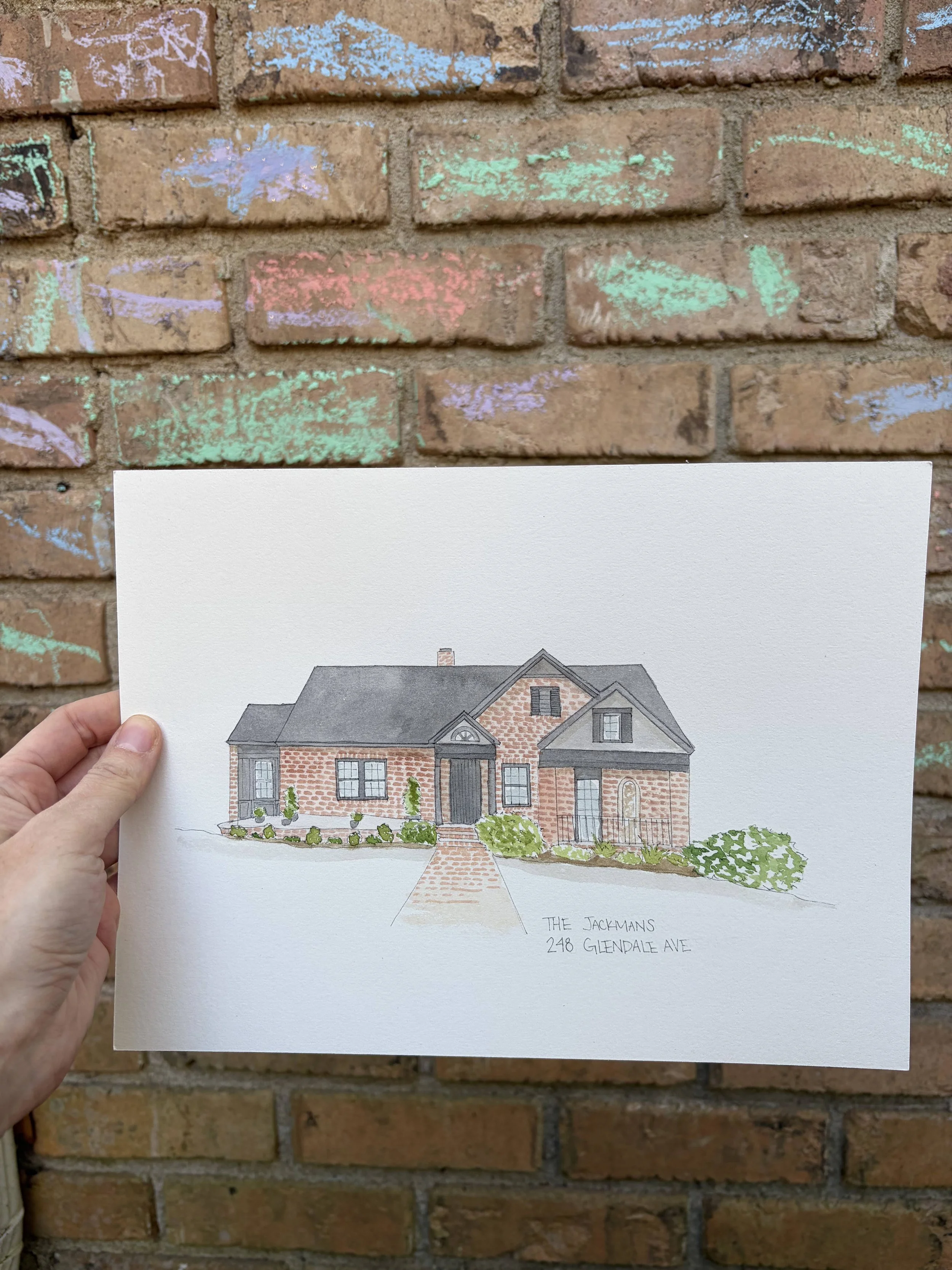Colorful drawing of a house with a brick pathway leading to the front door and the address '248 Glendale Ave' written below, held in front of a brick wall with chalk marks.