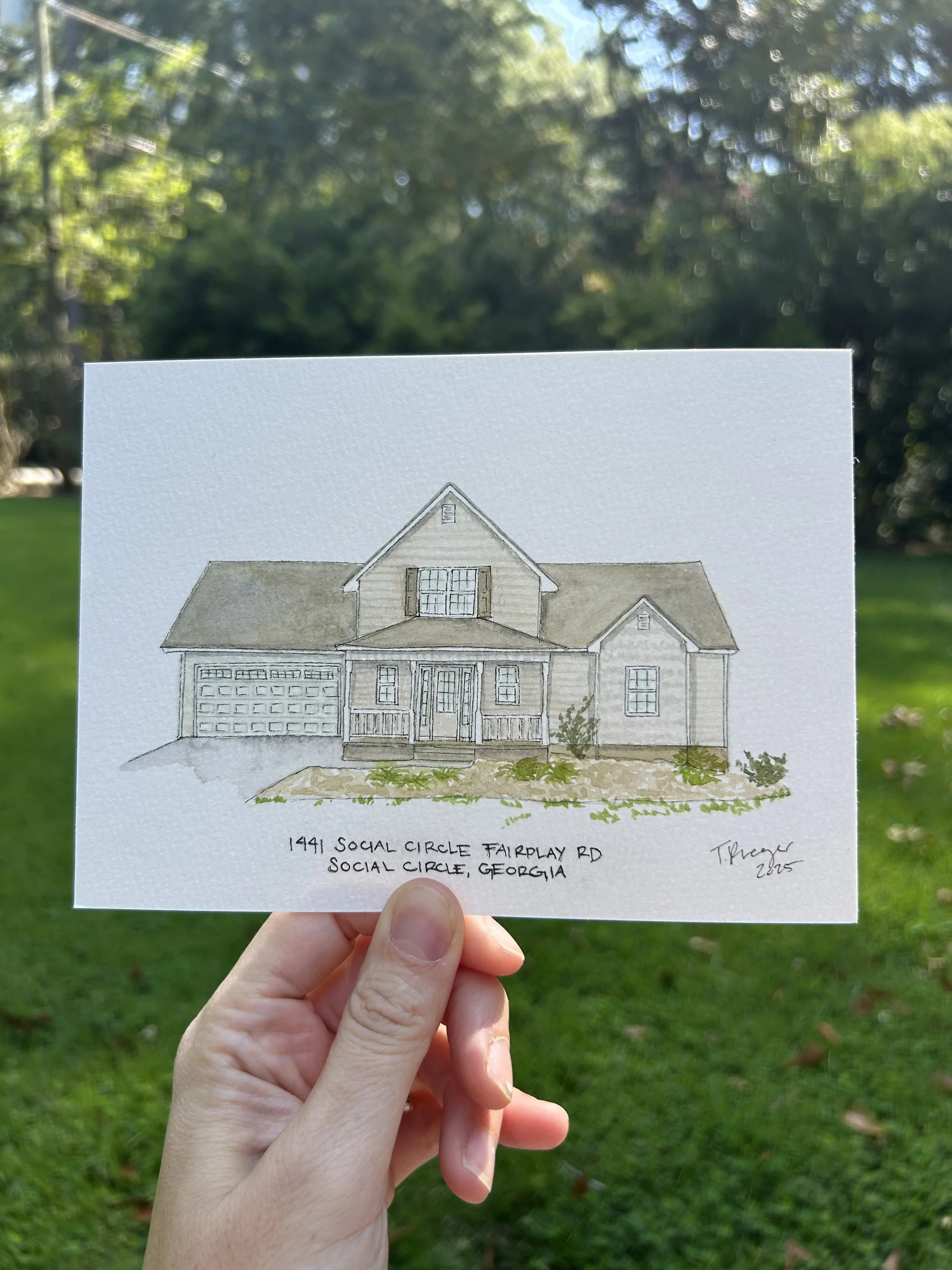 A person holding a watercolor illustration of a house with a gabled roof, a garage, and a porch in front of a grassy yard. The illustration is labeled with the address '1441 Social Circle Fairplay Rd, Social Circle, Georgia' and is signed 'T. Henry 2023'.