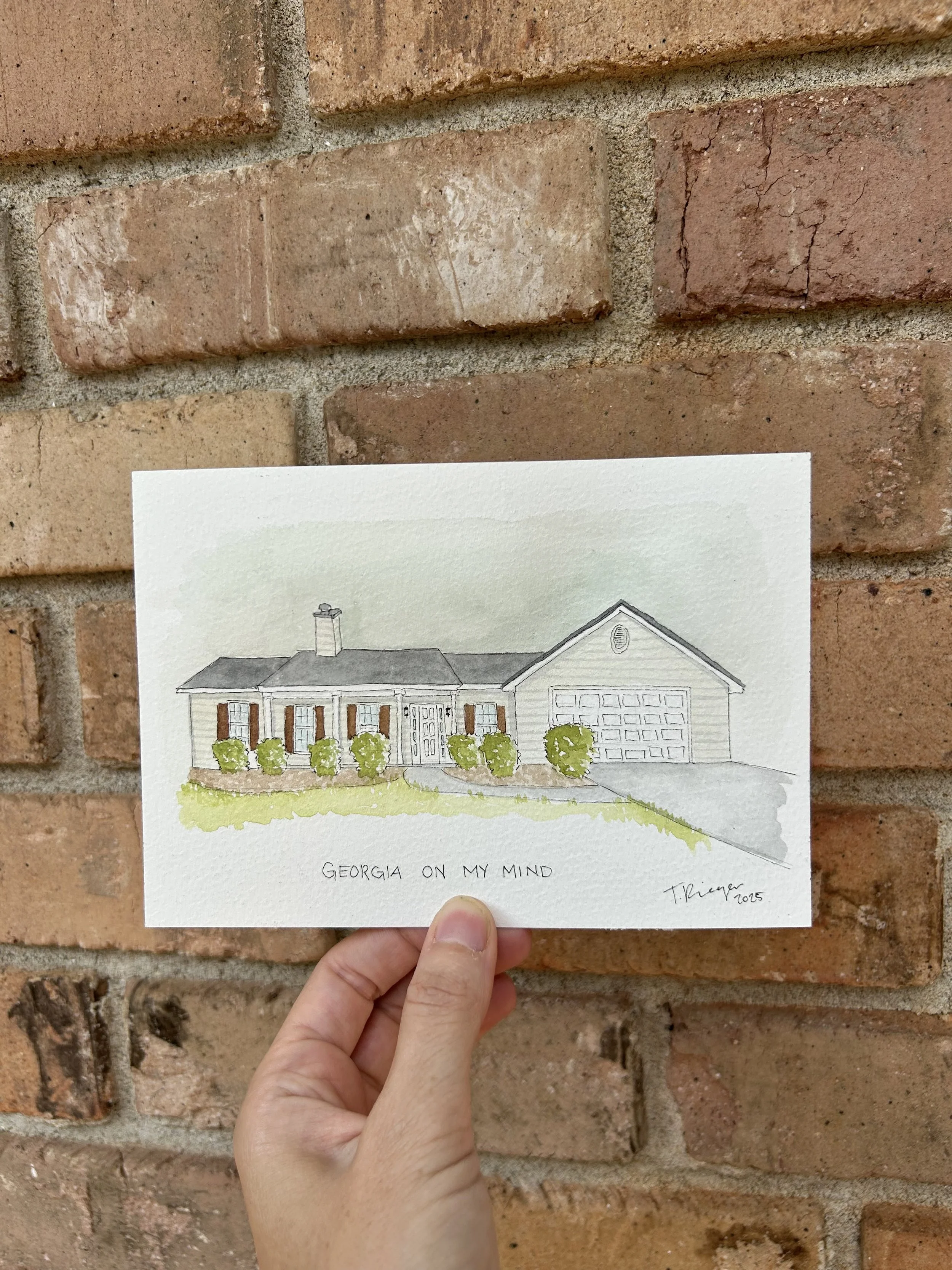 A person holding a drawing of a house in front of a brick wall. The drawing is labeled "Georgia On My Mind" and is signed by T. Krieger, 2015.