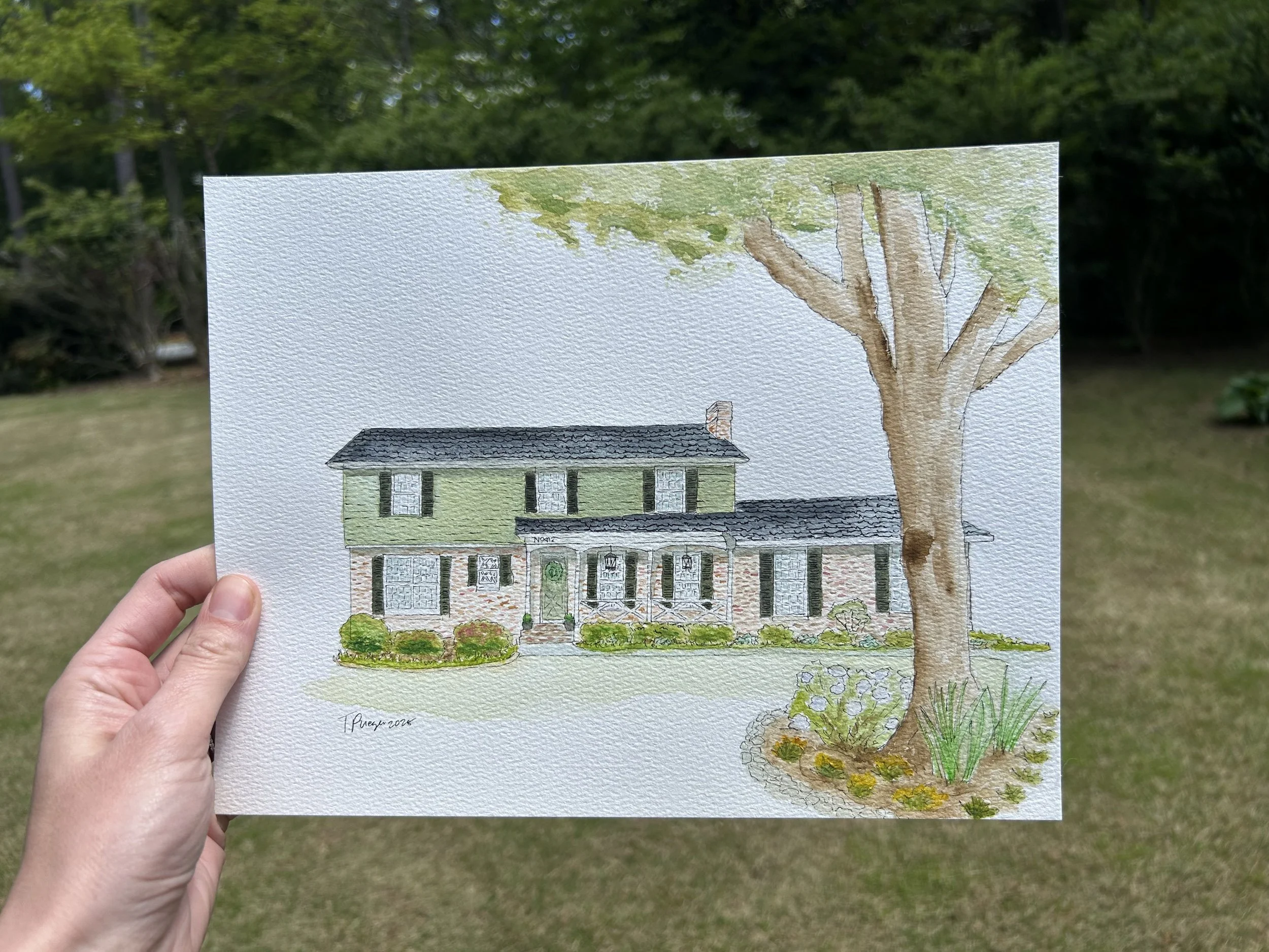 Watercolor painting of a two-story green house with a porch, surrounded by a garden, with large trees in the background, held by a person's hand outdoors.