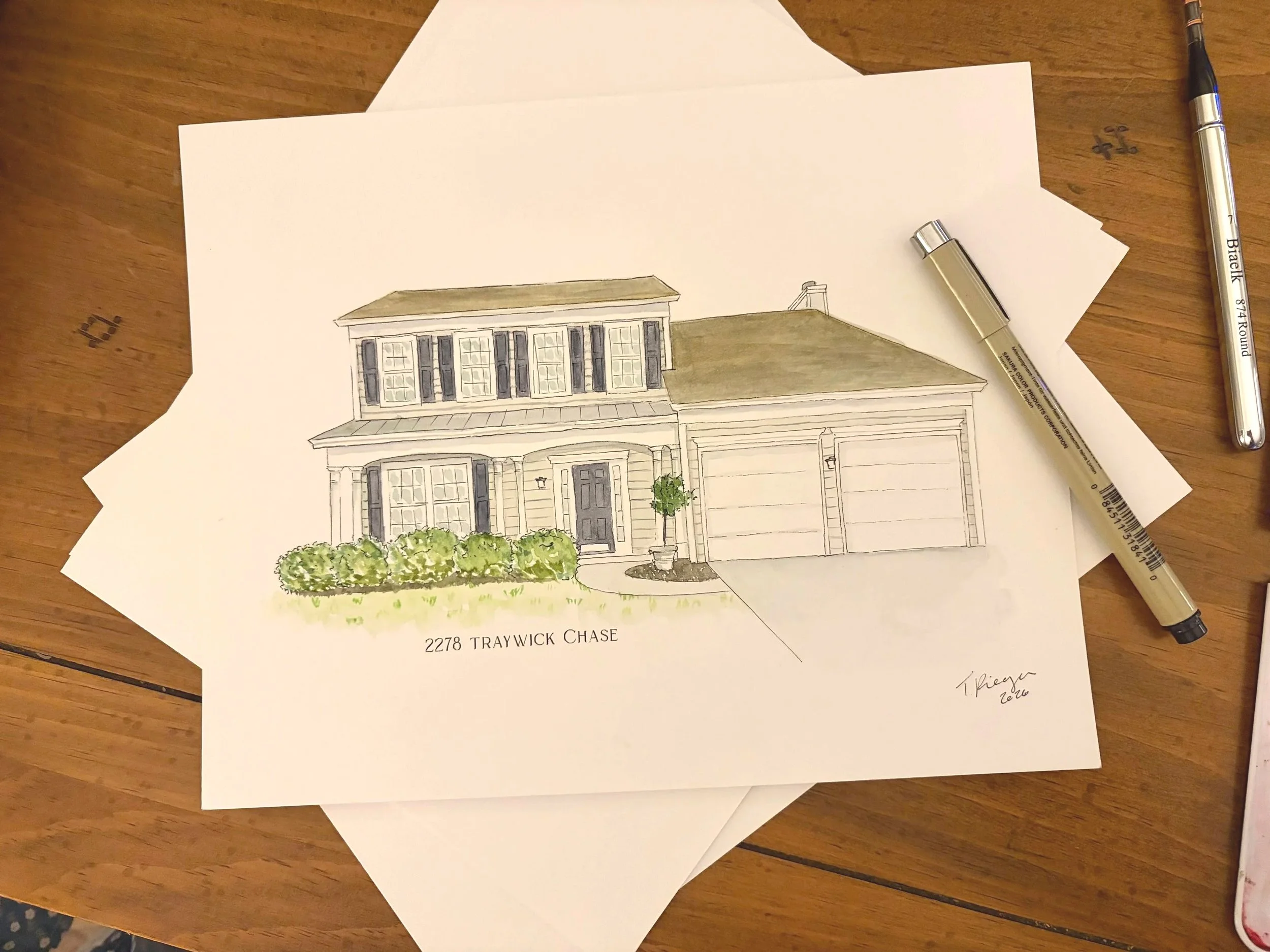 A hand-drawn architectural illustration of a house with a front porch, surrounded by bushes, on a piece of paper labeled "2278 TRAYWICK CHASE". Two pens are placed on the paper, one near the top right corner and the other near the middle right side, on a wooden surface.